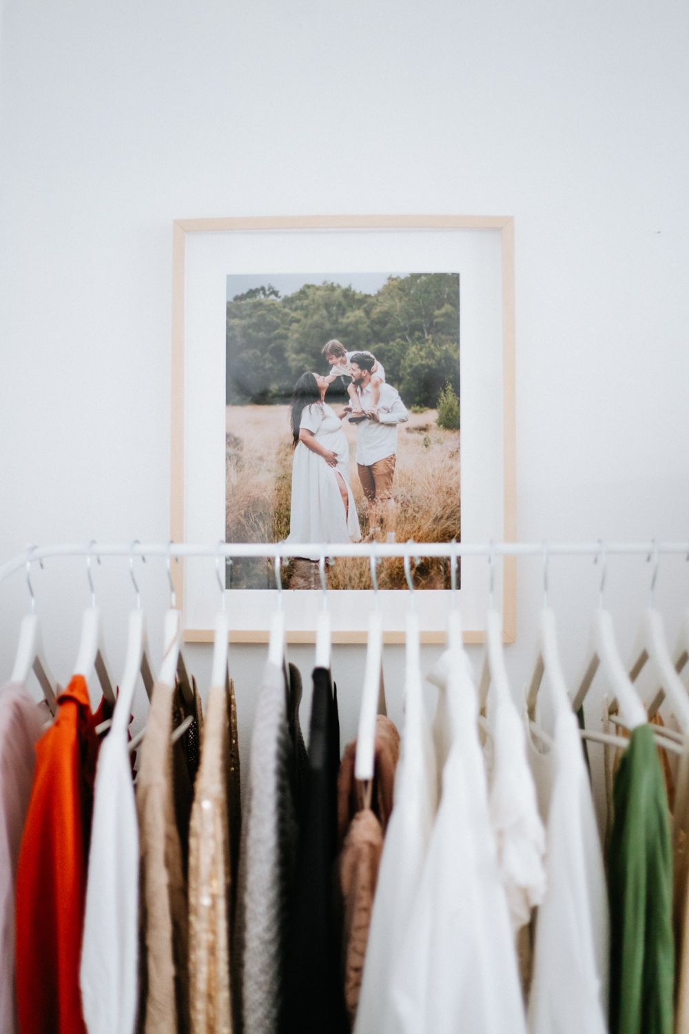 A framed photo sits above a closet rack filled with clothing in various colors from white to green.