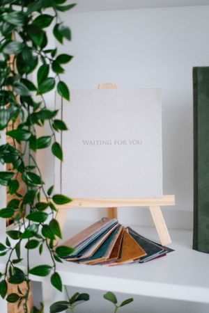 Minimalist white card reading 'waiting for you' on wooden easel with fabric swatches and green plant decor.