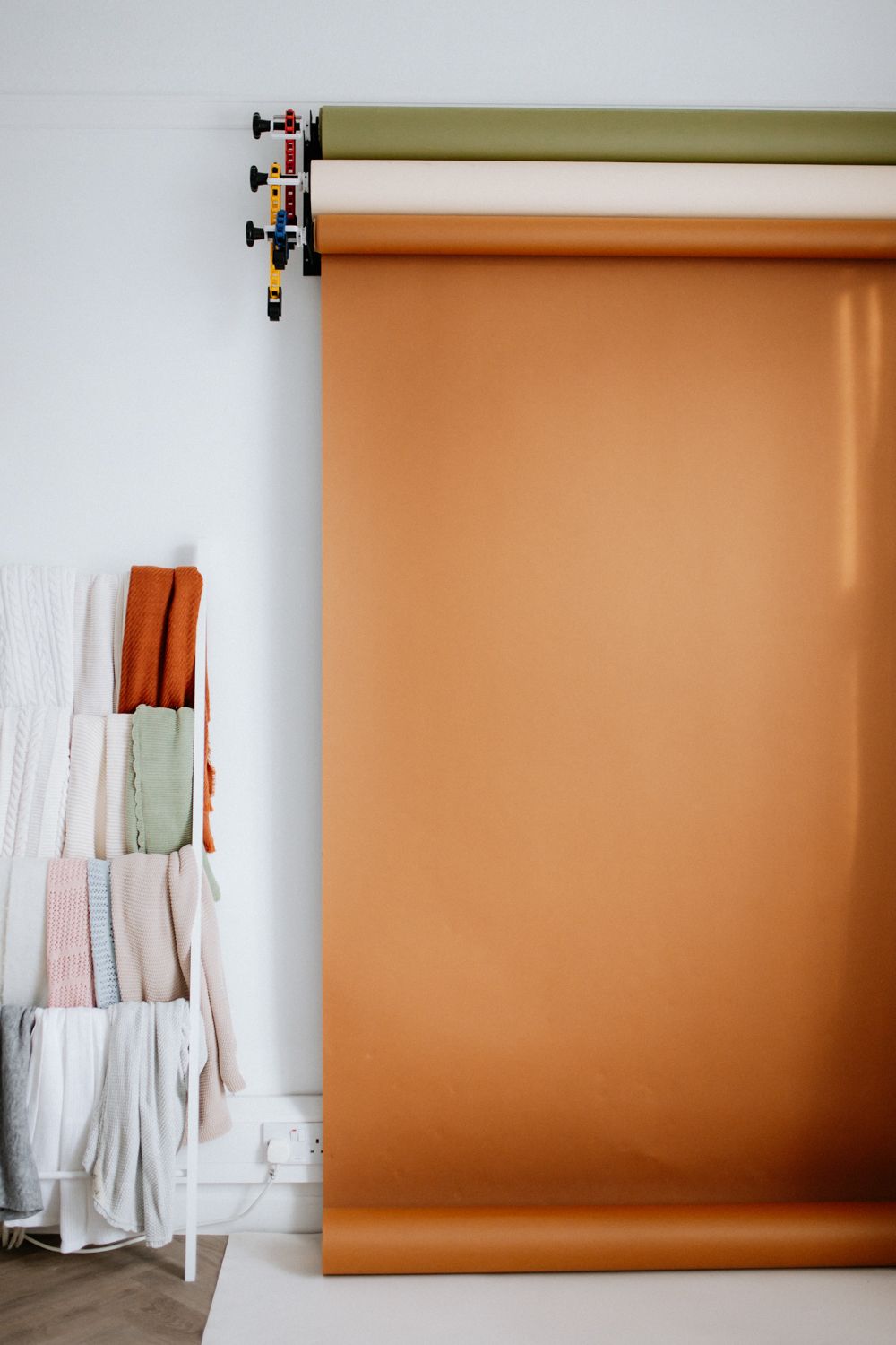 Photography studio setup with rolled orange backdrop paper and stack of folded fabrics in muted tones against white wall.