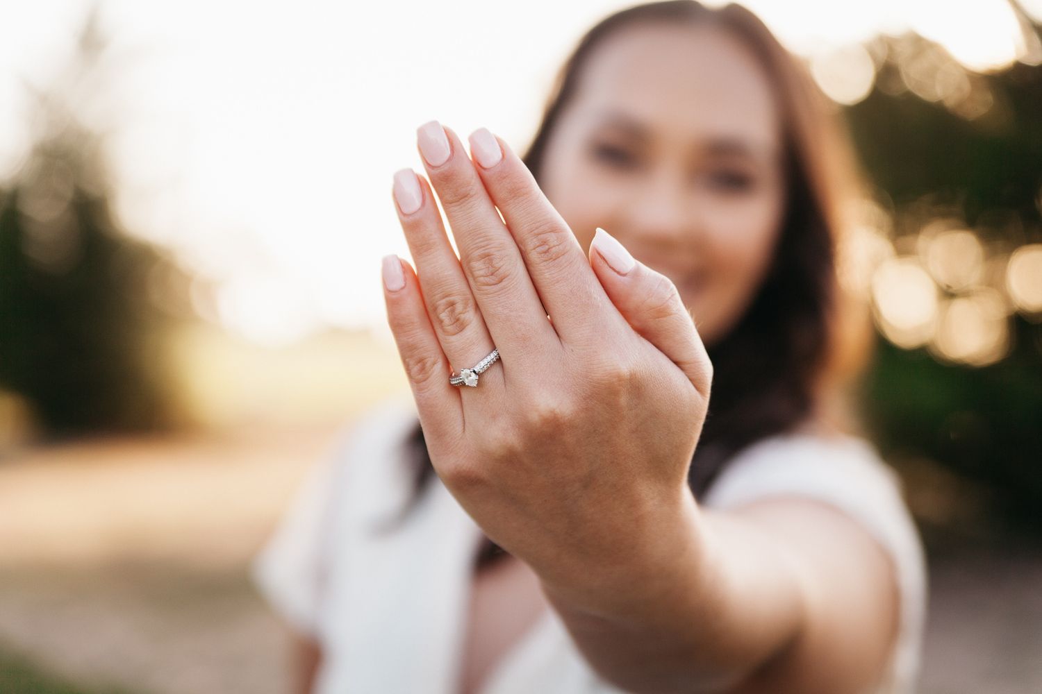 A woman holds up her left hand to show a diamond engagement ring at sunset.