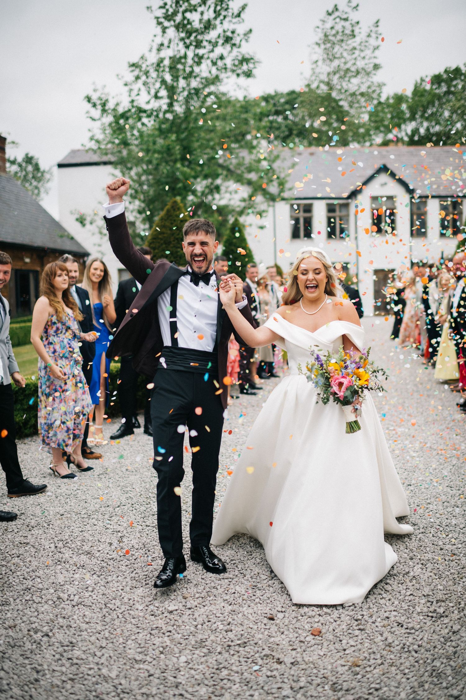 A wedding dress train flows elegantly across dark gravel as guests gather for an outdoor celebration. Editorial wedding photographer.