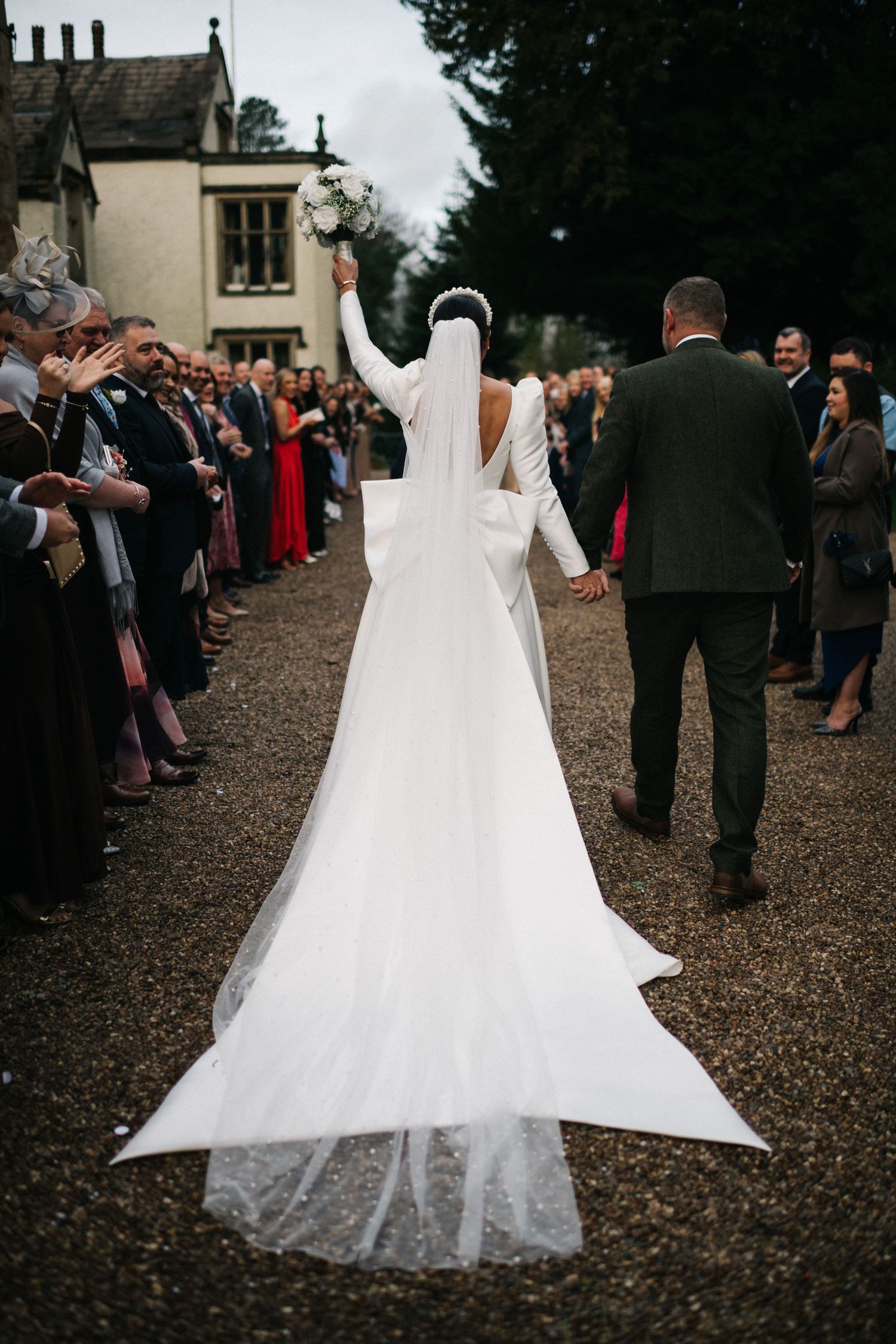 A wedding dress train flows elegantly across dark gravel as guests gather for an outdoor celebration. Editorial wedding photographer.