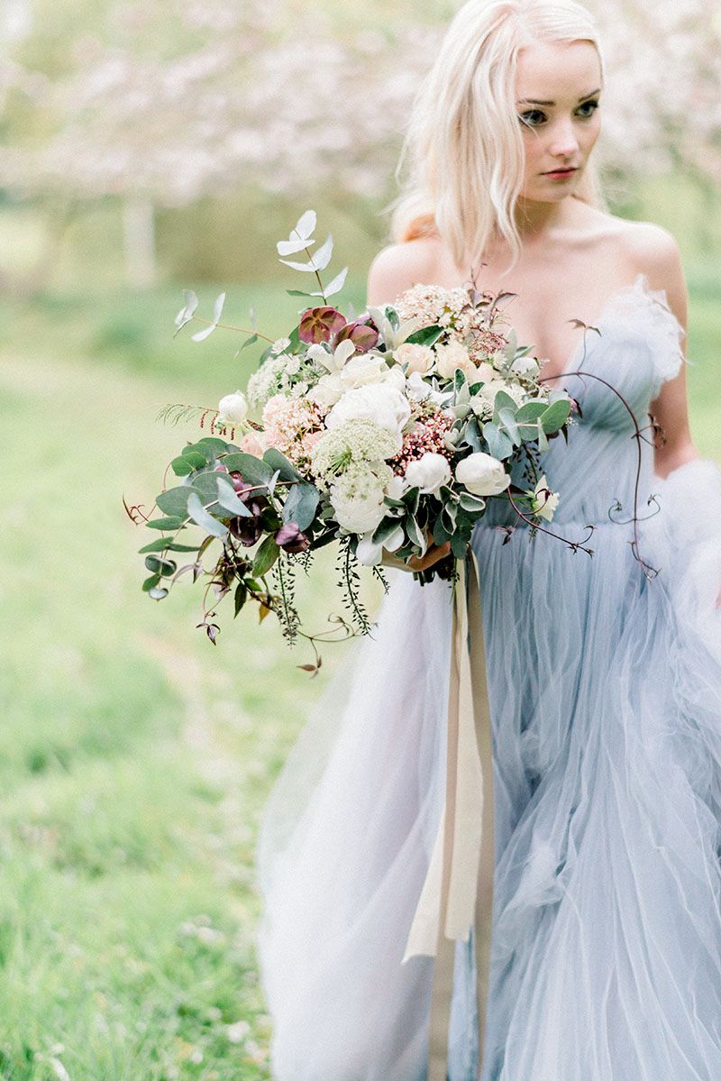 Blonde bride in off-shoulder dusty blue tulle gown holding lush ivory and burgundy bouquet with eucalyptus in garden.