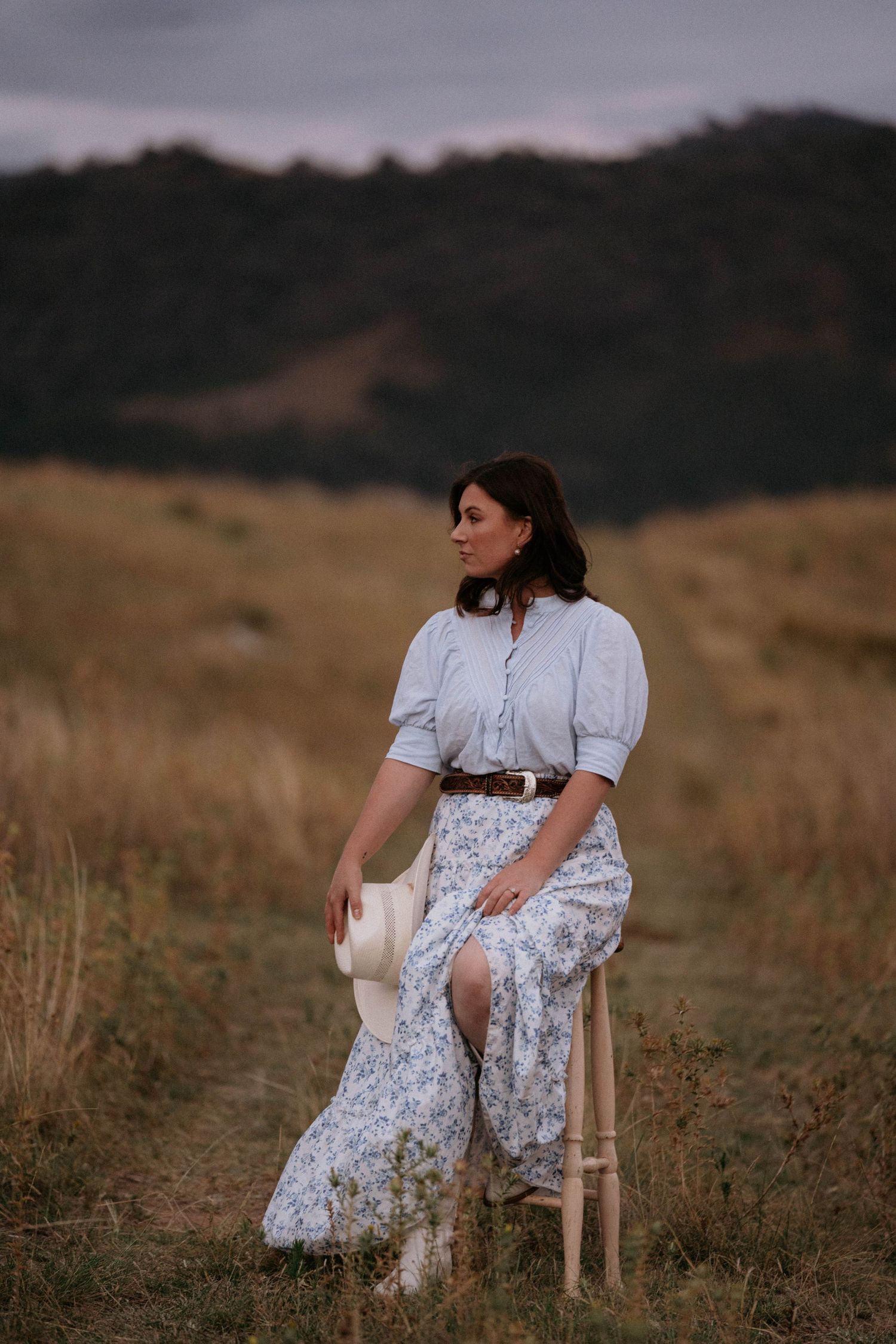 A woman in a vintage-style outfit with a floral skirt and white blouse sits on a stool in a golden meadow at dusk.