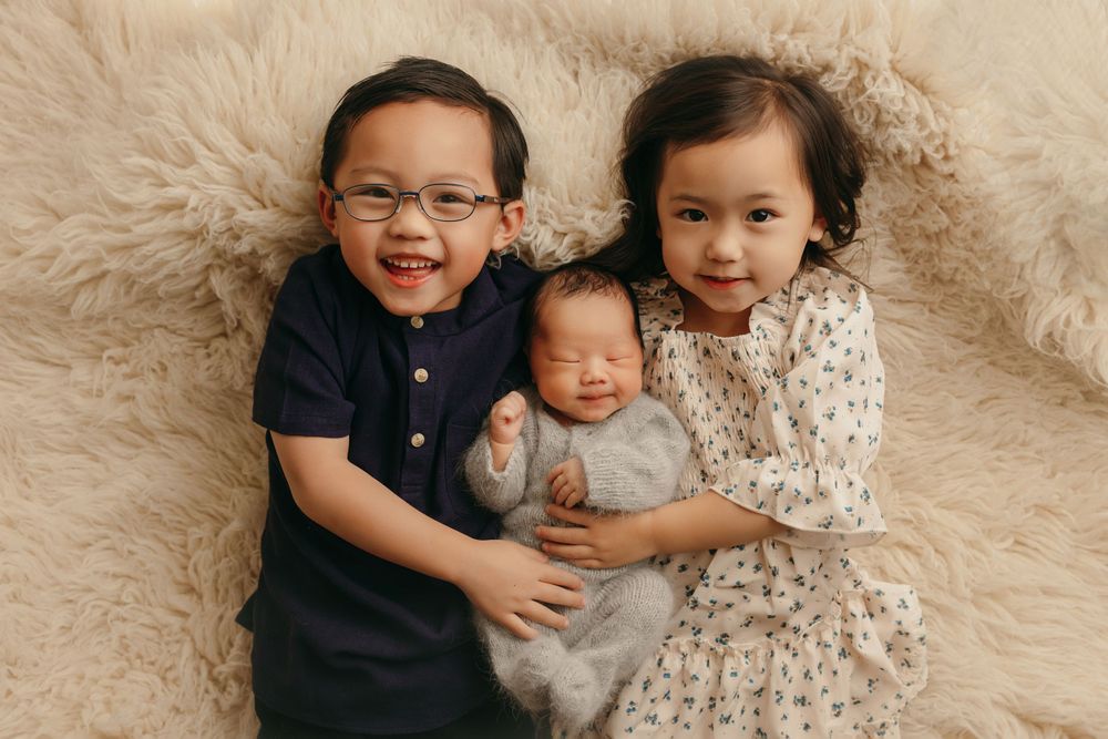 Three siblings pose together on a beige furry blanket, with the eldest two holding their newborn sibling.