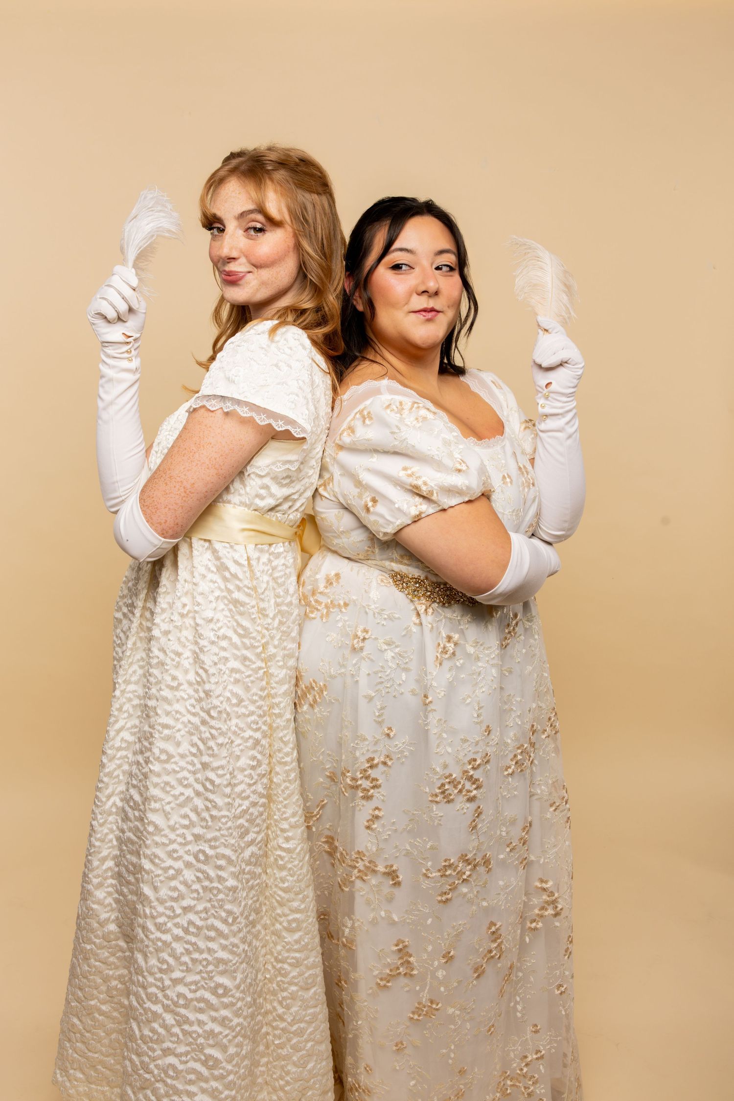 Two women in elegant cream-colored regency-style dresses and white gloves pose back-to-back.