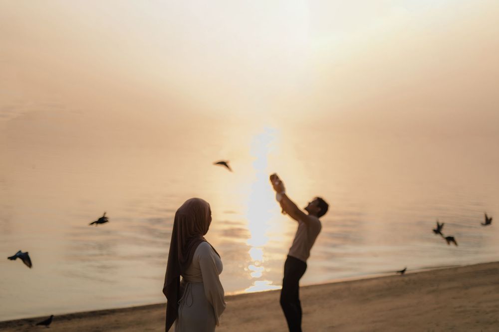 Couple on a misty beach at sunset, with birds flying over shimmering golden water as man reaches toward the sky.
