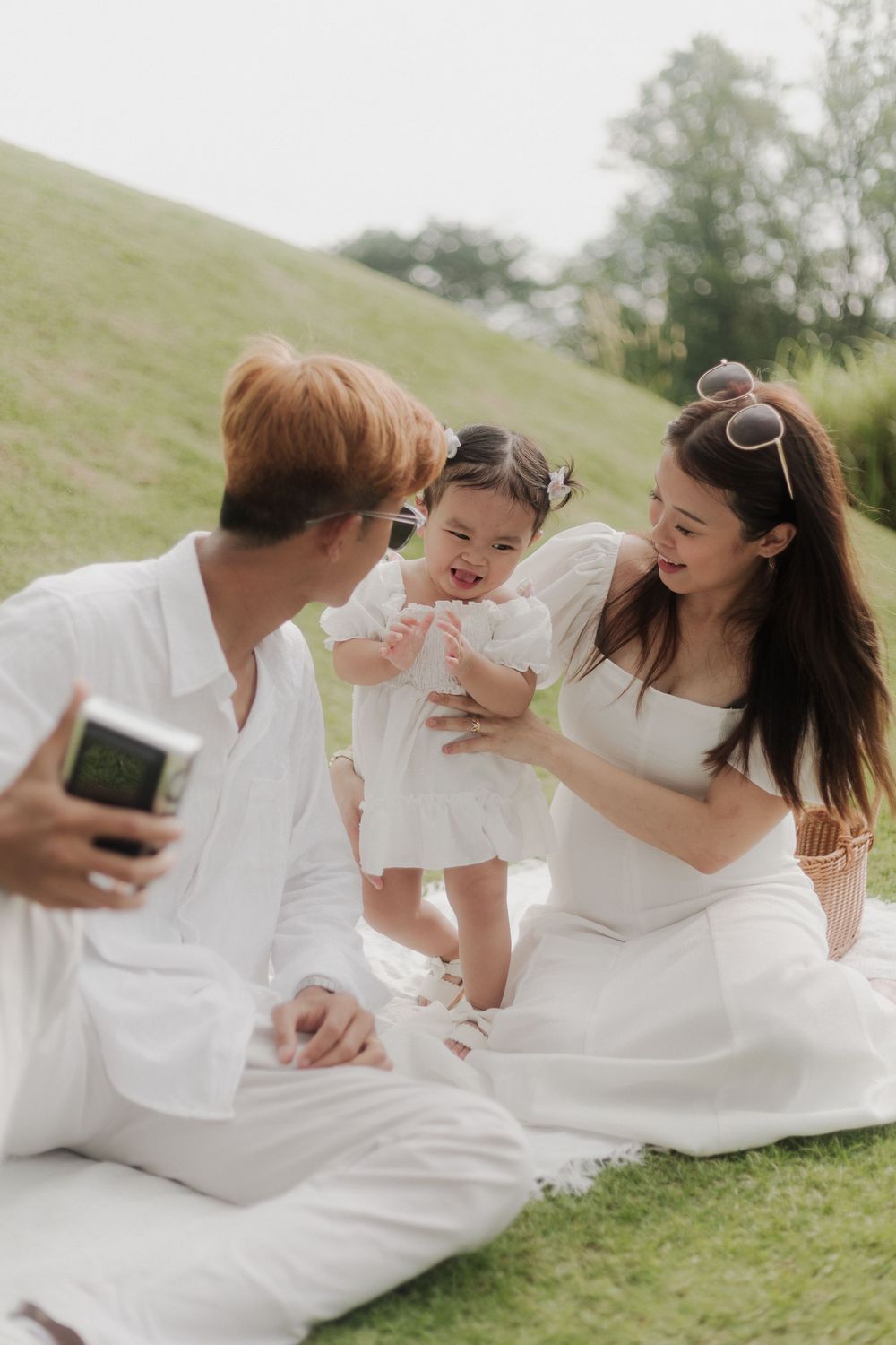 Family in white outfits enjoying a picnic on green grass, parents playing with their happy toddler daughter outdoors.