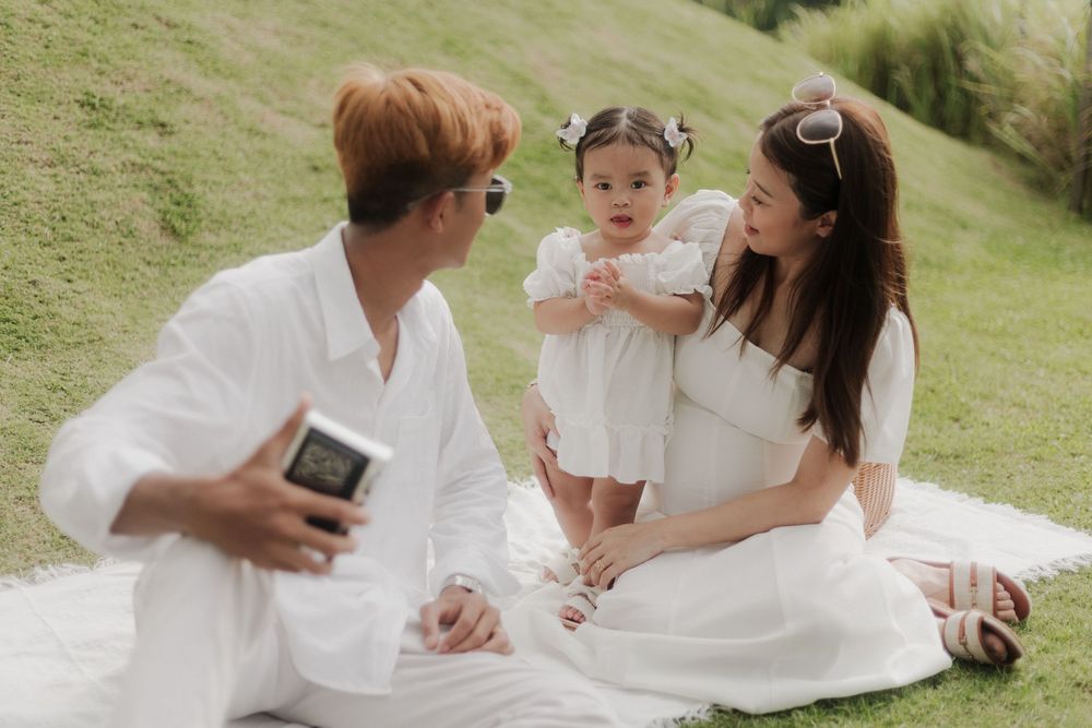 Family of three sitting on white blanket outdoors, parents in white outfits with toddler girl on sunny green lawn.
