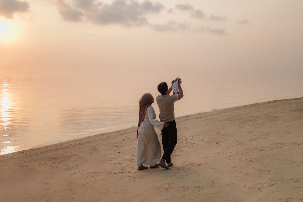 Couple dancing on sandy beach at sunset with calm ocean waters reflecting warm golden light in the background.