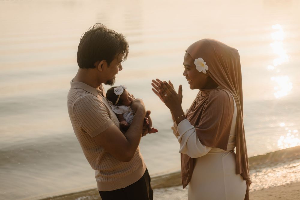 A couple in warm golden light by the water, with the man holding a newborn baby while the woman gestures lovingly.