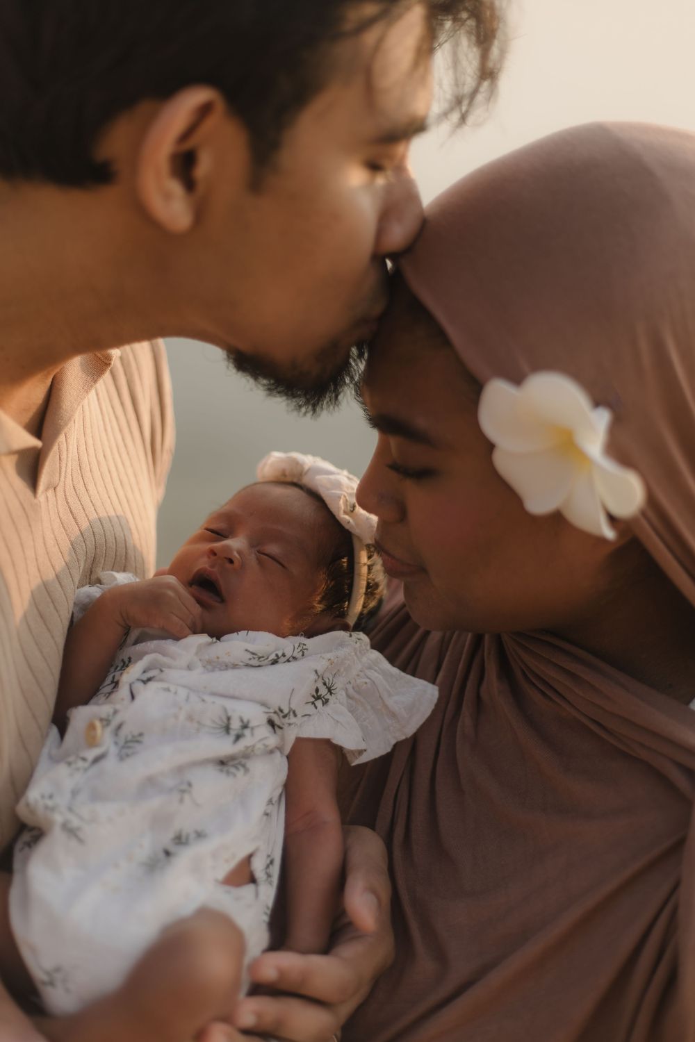 Muslim couple kissing over their newborn baby, mother wearing hijab with white flower, warm golden light family portrait.