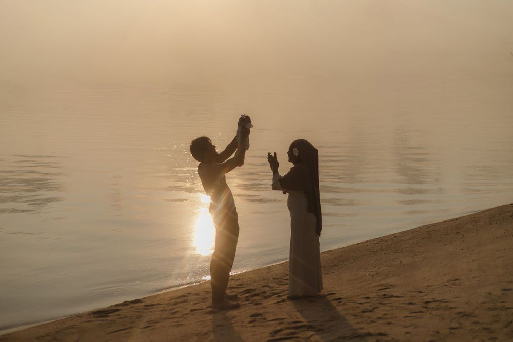 Two silhouettes play on a misty beach at sunset, one reaching up while the other gestures near a calm reflective shoreline.