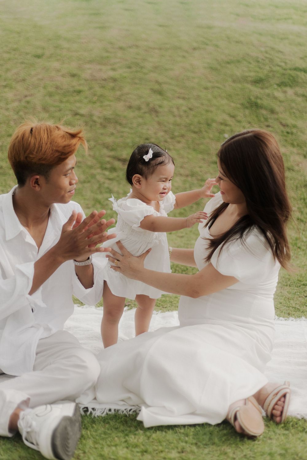 A happy Asian family in white outfits sits on grass, with parents helping their toddler daughter stand between them.