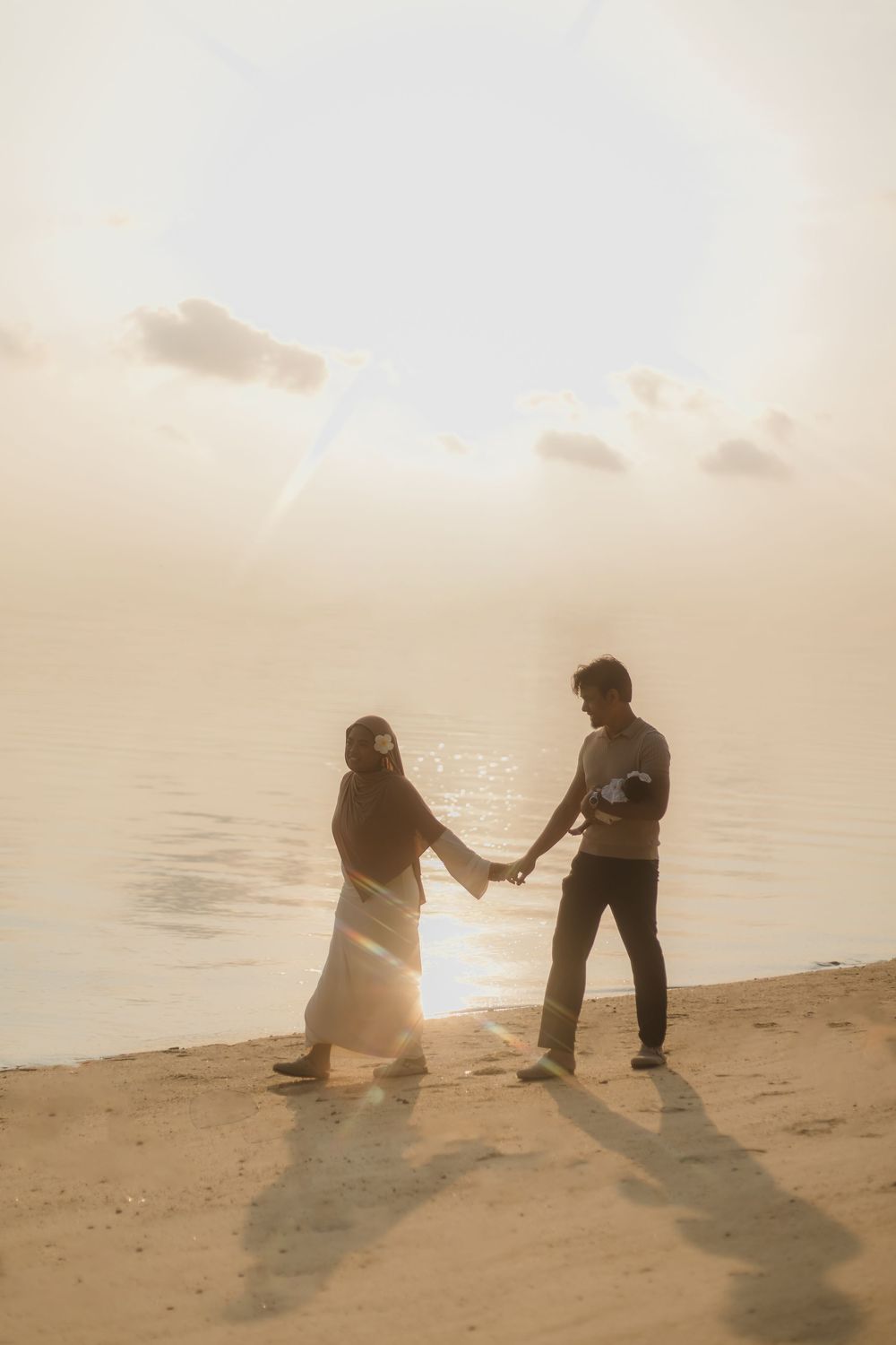 Couple holding hands on a sandy beach at golden sunset with warm lens flare and shimmering water in the background.