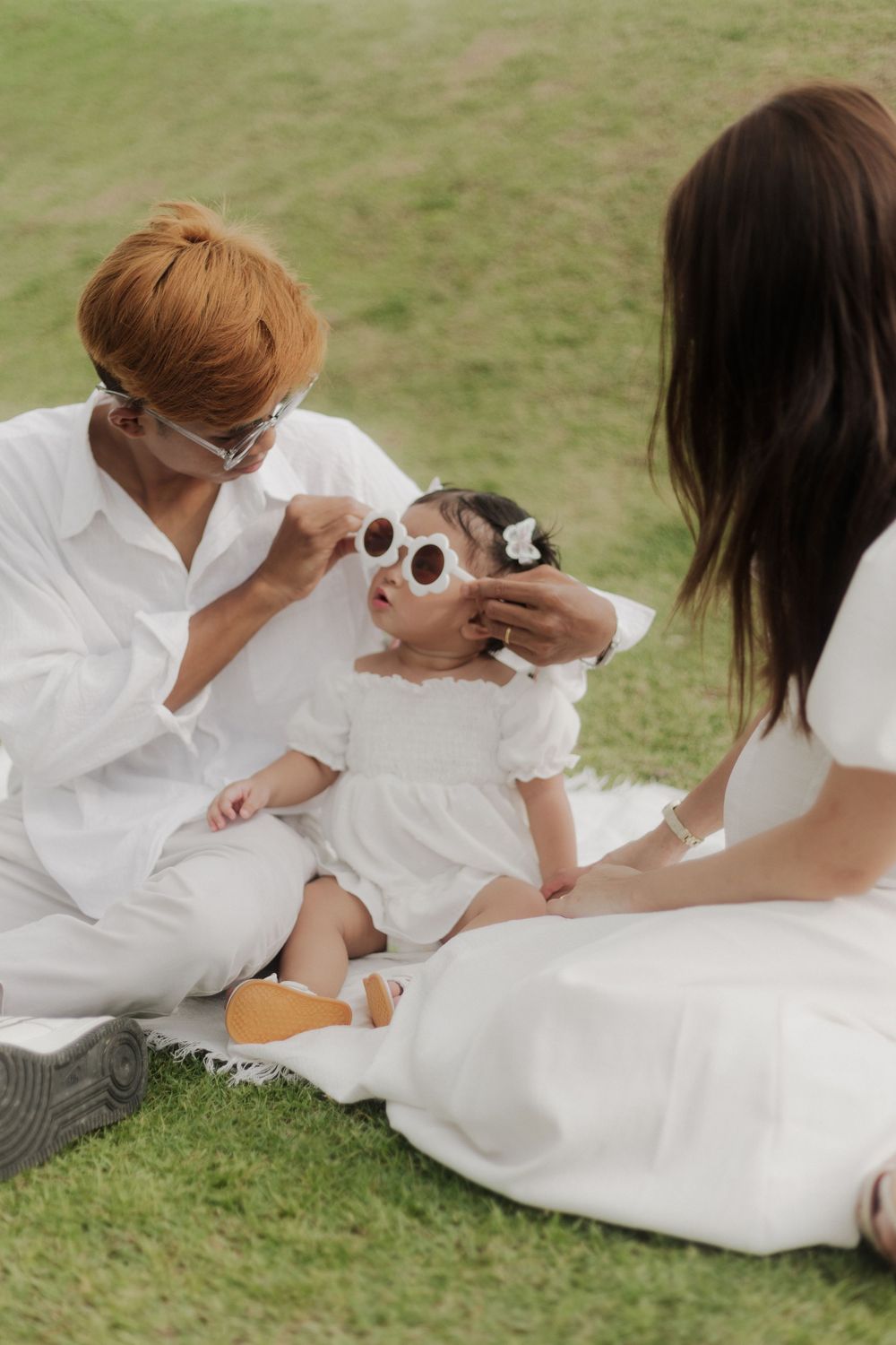 Family in white outfits sitting on grass, parents putting sunglasses on cute baby girl during outdoor picnic.