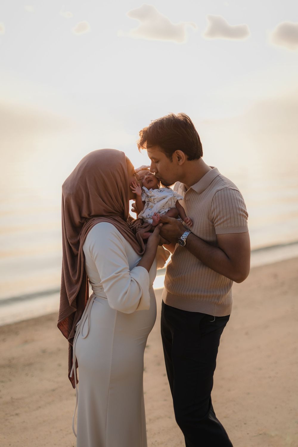 Muslim couple kissing their newborn baby on a sandy beach at golden sunset hour.