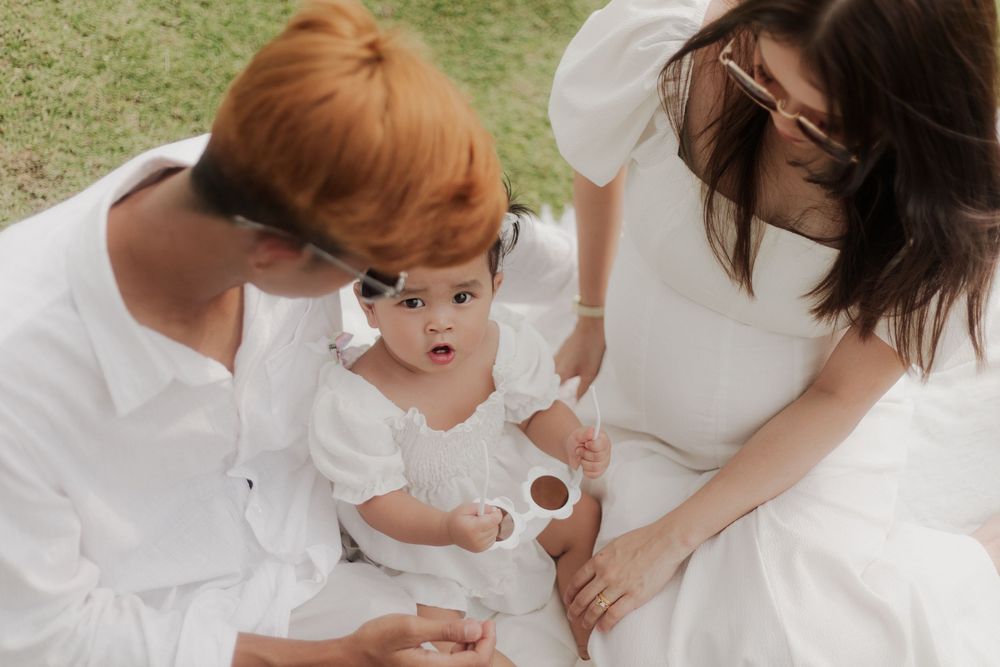 Two parents in white outfits sit outdoors on grass with their baby girl dressed in white, sharing a tender family moment.