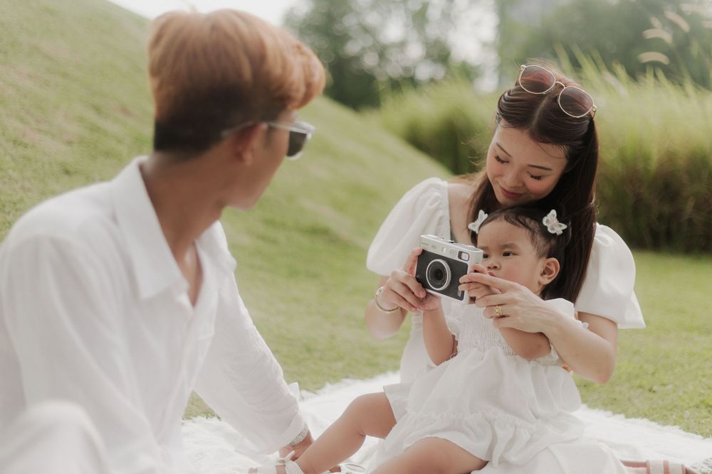 Family sitting on grass, mother showing baby a vintage camera while father watches, all wearing white outfits outdoors.
