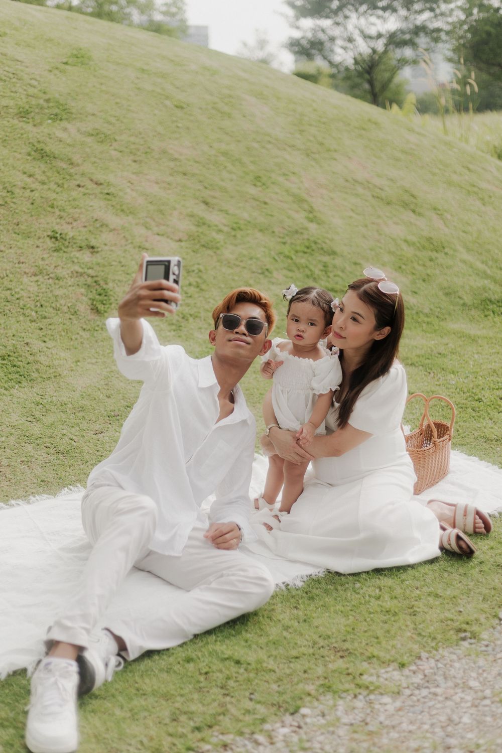 Asian family in matching white outfits taking selfie with camera while sitting on blanket in green park.