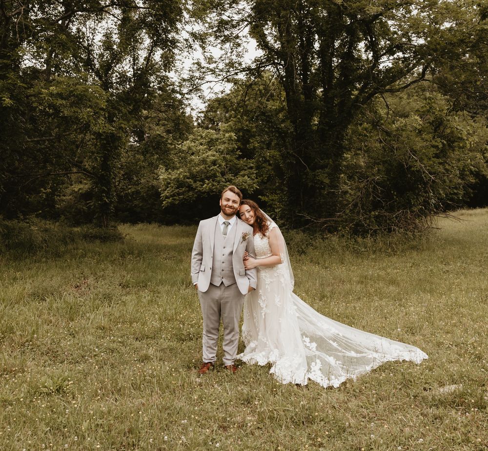 A couple stands together in a rustic outdoor setting wearing wedding attire on their special day.
