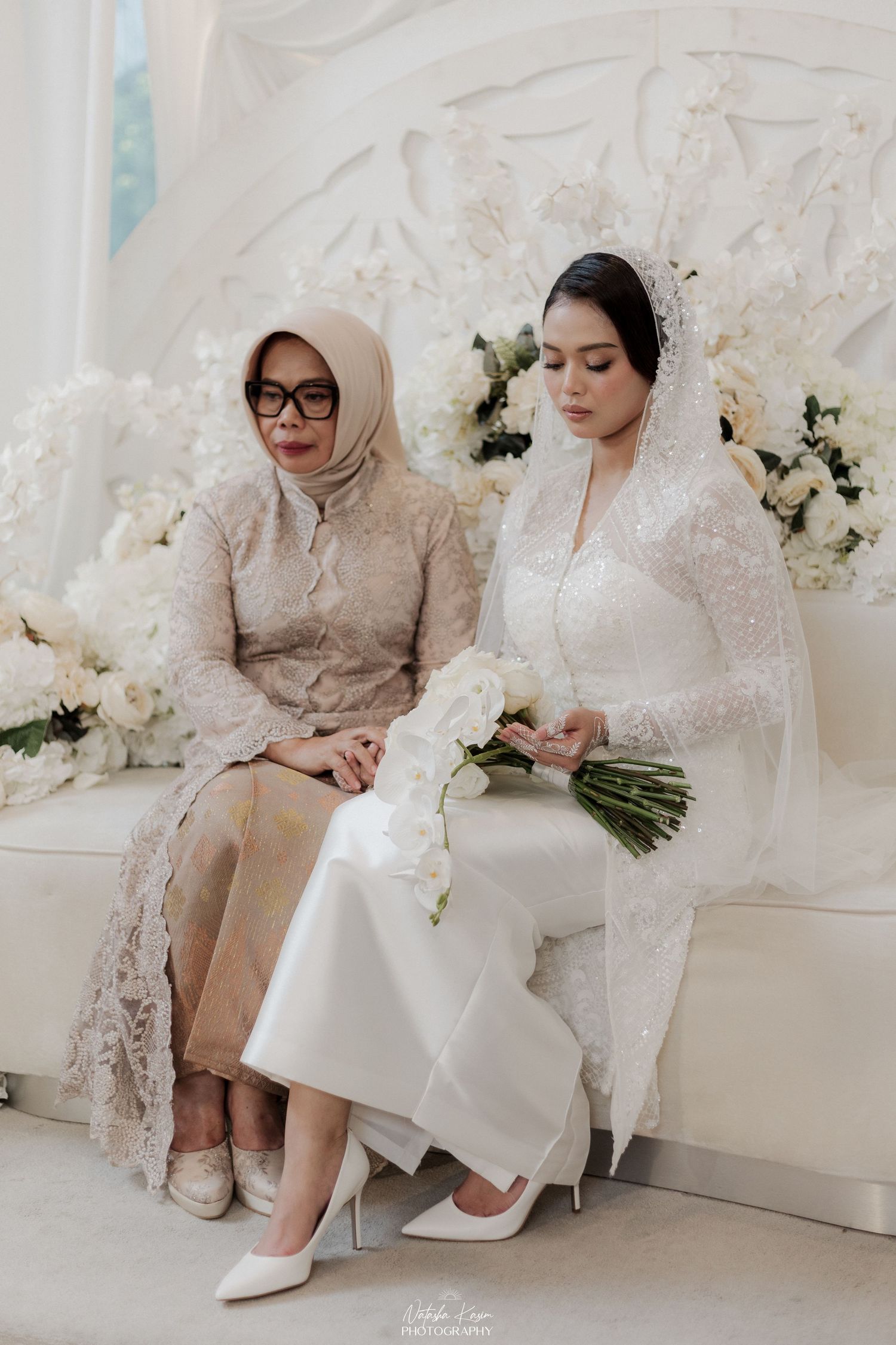 Two women in elegant white and beige attire sit together in an elegant white room with floral decor.