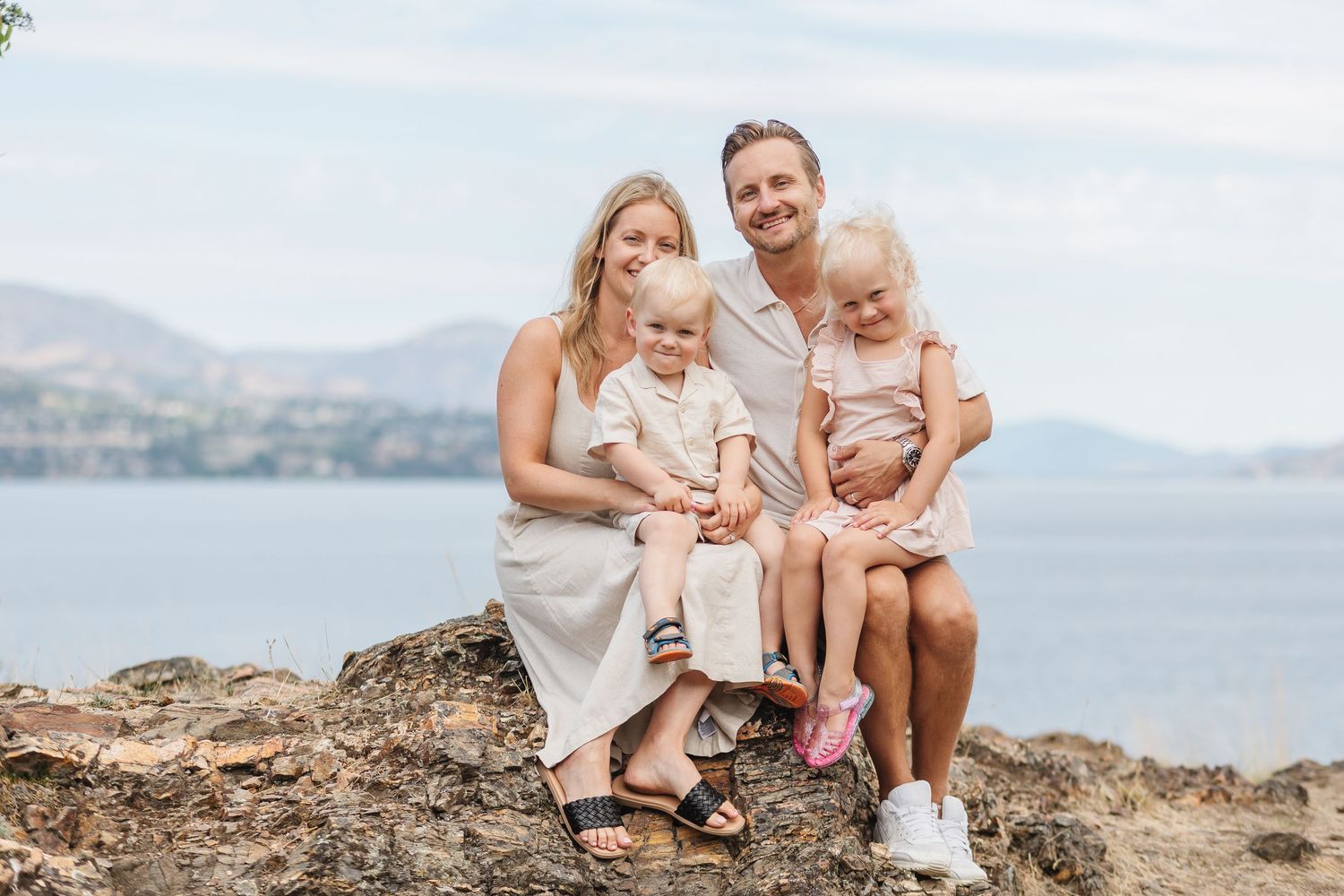 A family sits together on coastal rocks overlooking a scenic mountain and water landscape during sunset.