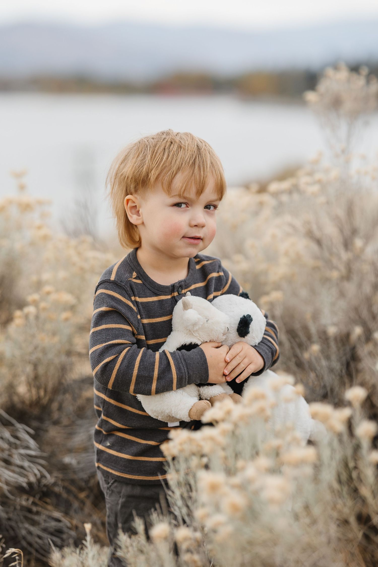 A small child in a striped sweater plays with a white plush toy among dried brush in a natural outdoor setting.