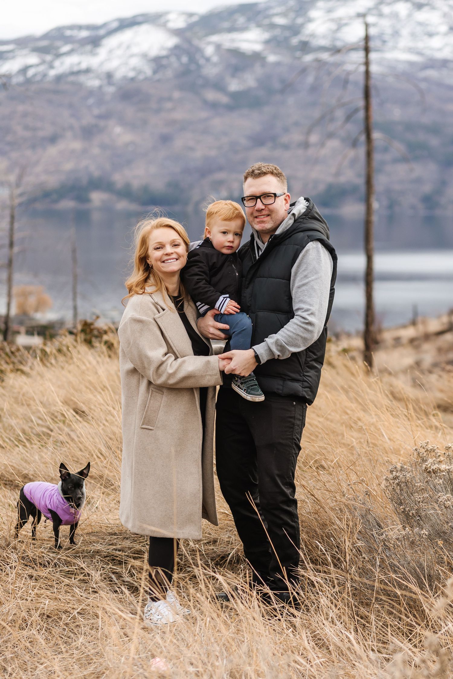 Family poses together on hillside with mountains and lake in background, accompanied by small dog for family photos in West Kelowna