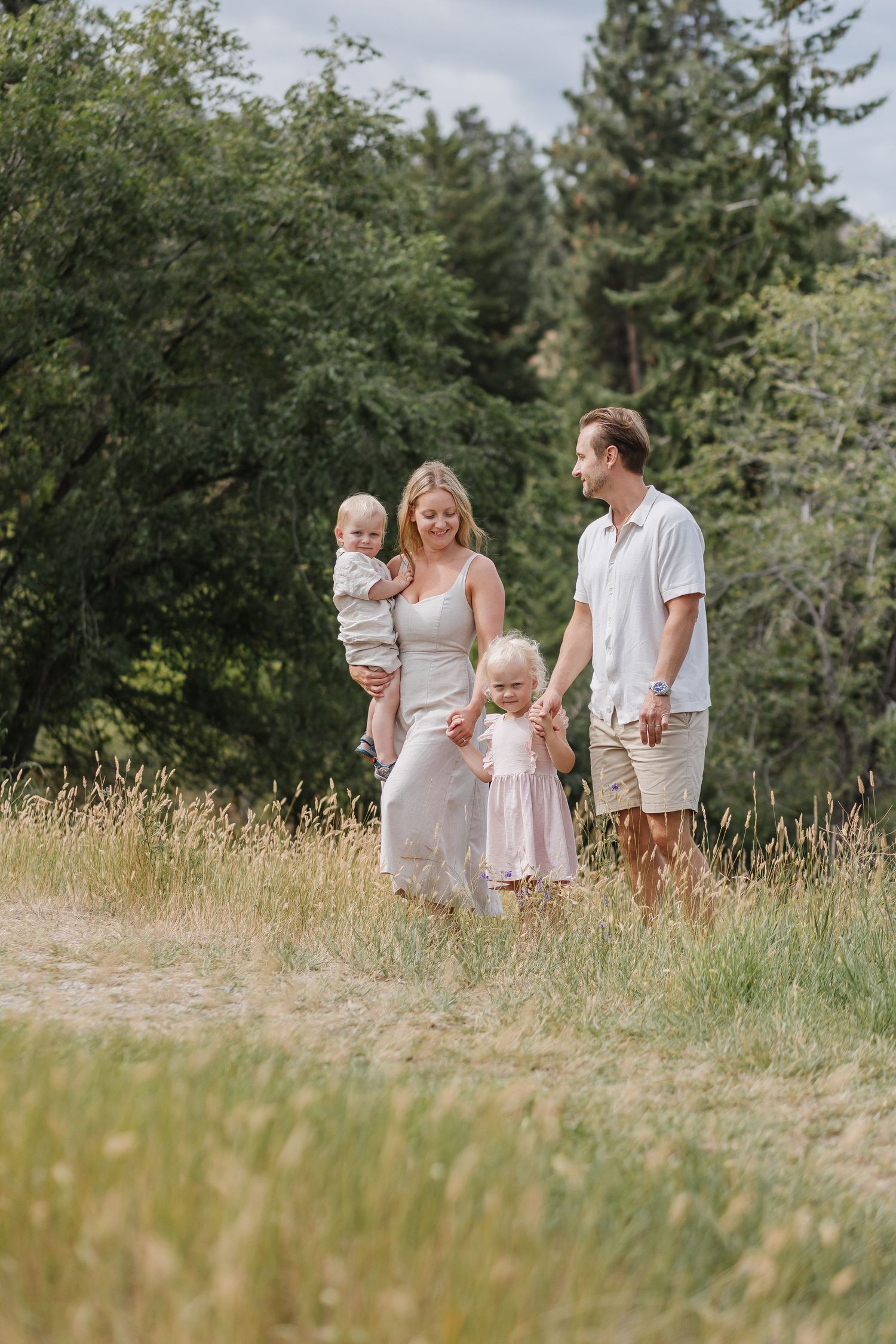 A family in white outfits walks together through a sunny meadow with tall grass and trees in the background.