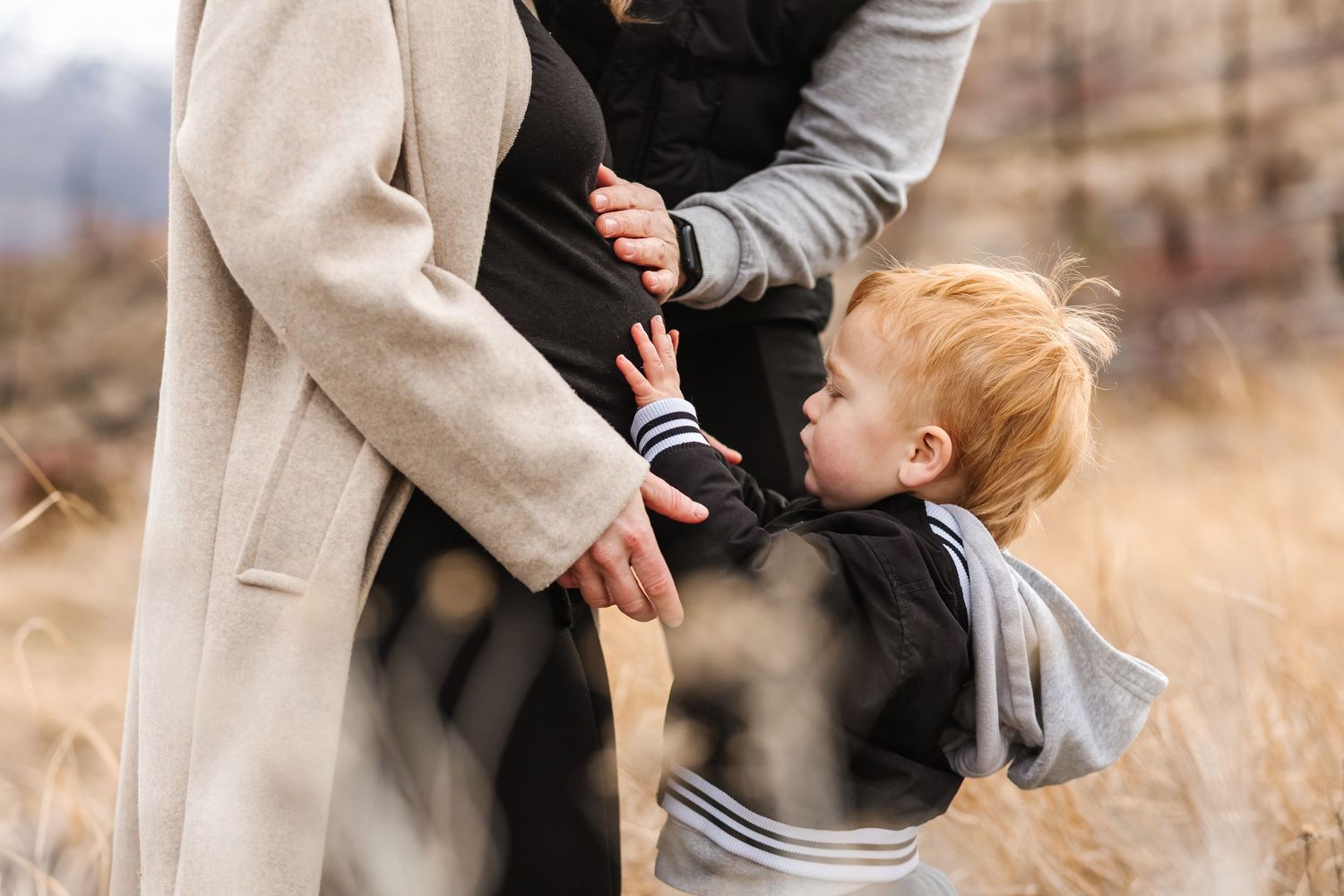 Candid family moment captured in golden field with mountains in background of pregnancy maternity photoshoot in West Kelowna