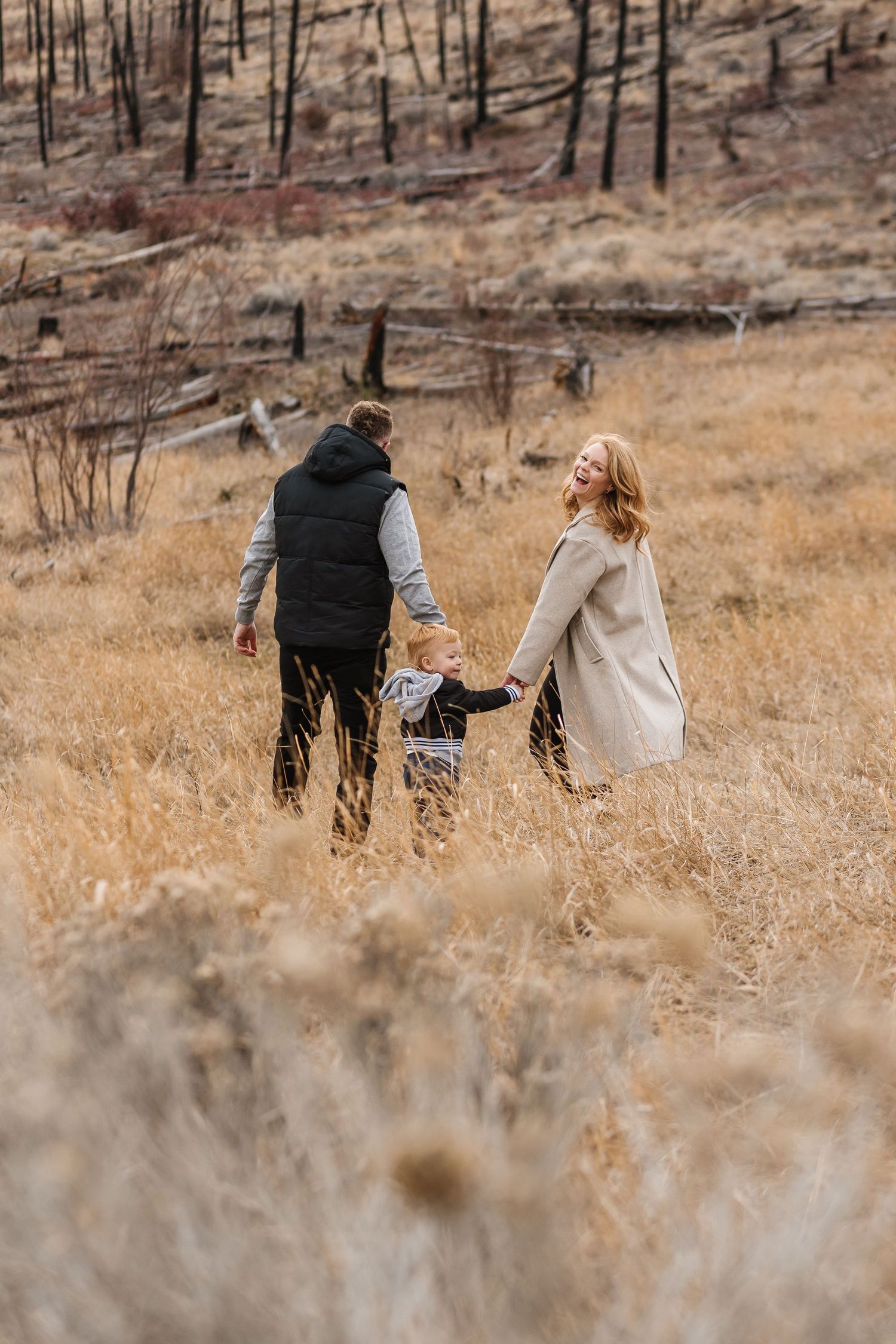 Couple walking through tall grass with burnt trees on hillside behind them for a family photoshoot in west Kelowna