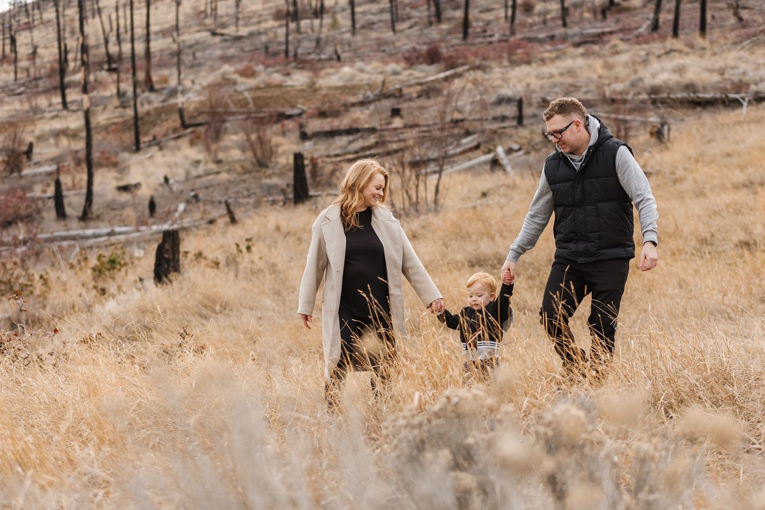 Couple holding hands while walking through golden autumn field with burnt forest in background in West Kelowna for family photos