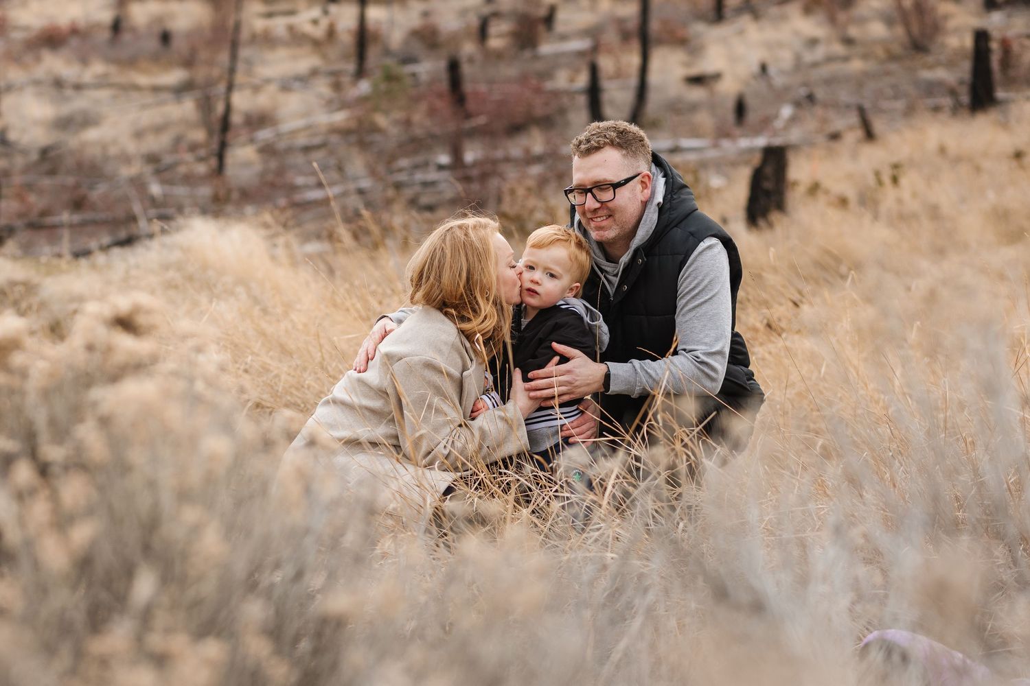 Intimate family moment captured in tall grass with burnt hillside in background in West Kelowna