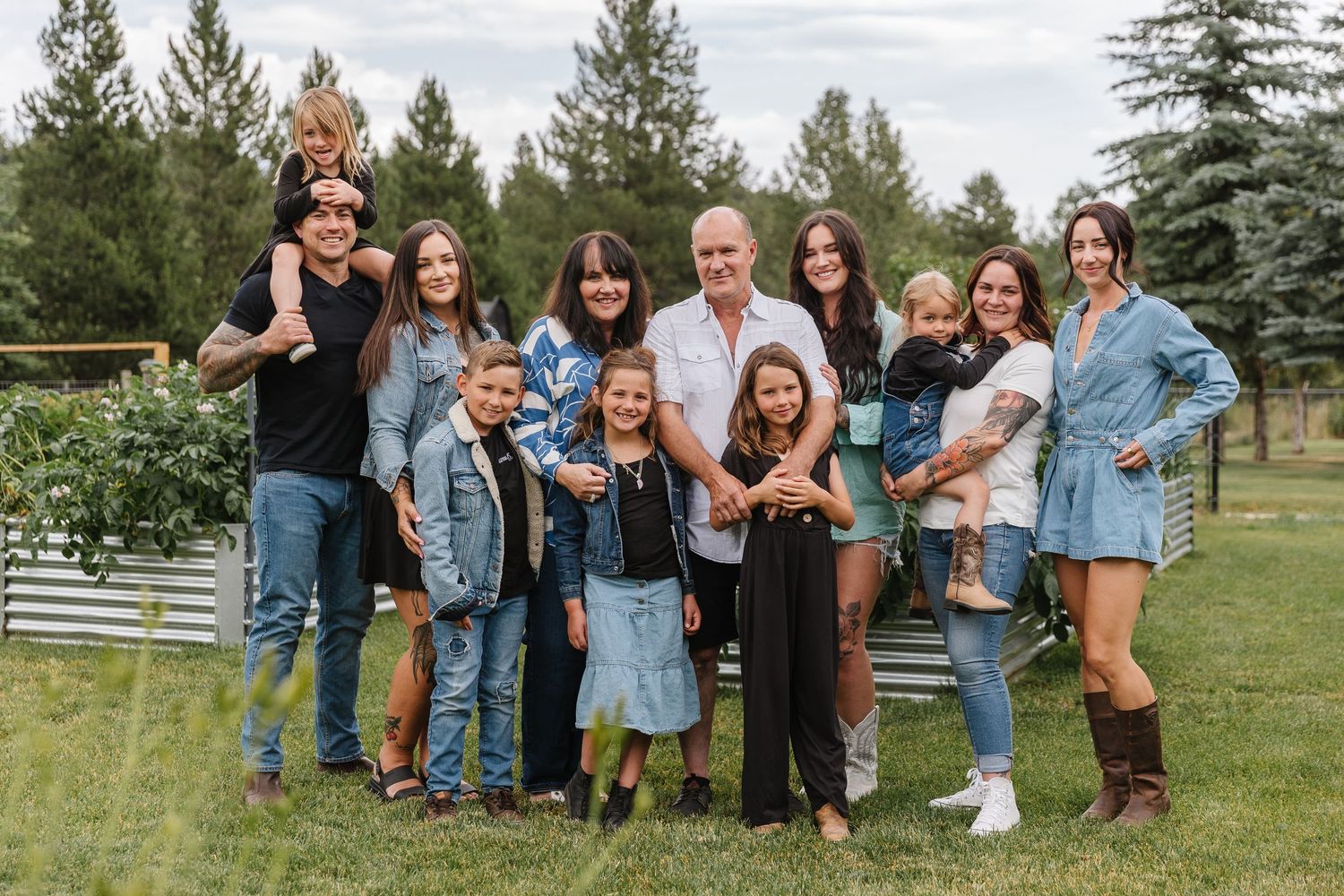 A group of people wearing denim and casual attire pose together outdoors in a grassy area with evergreen trees.