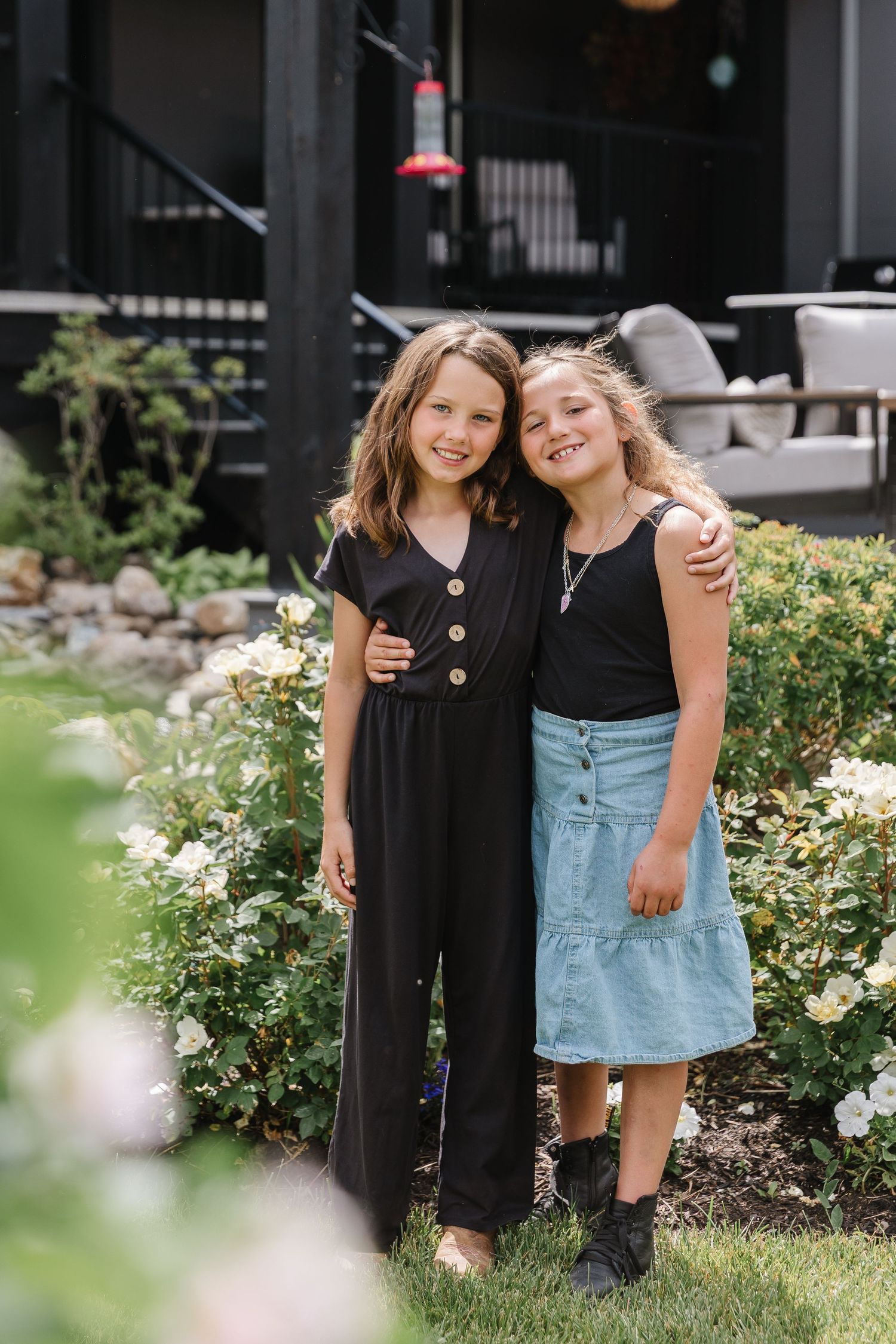 Two young friends pose together in a garden with blooming flowers, one wearing a black jumpsuit and one in denim.