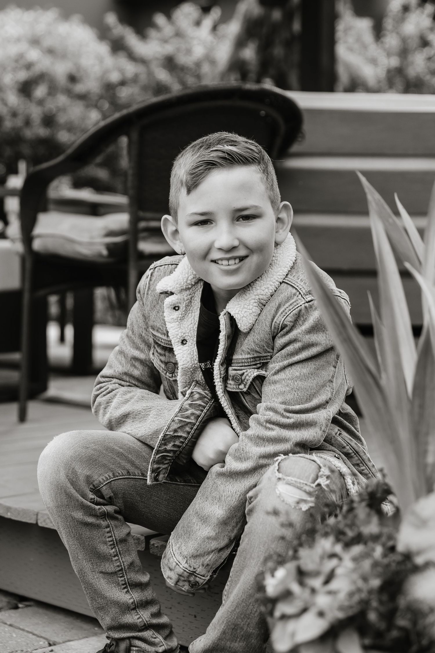 A young child in denim and a gray sweater poses playfully on outdoor furniture.