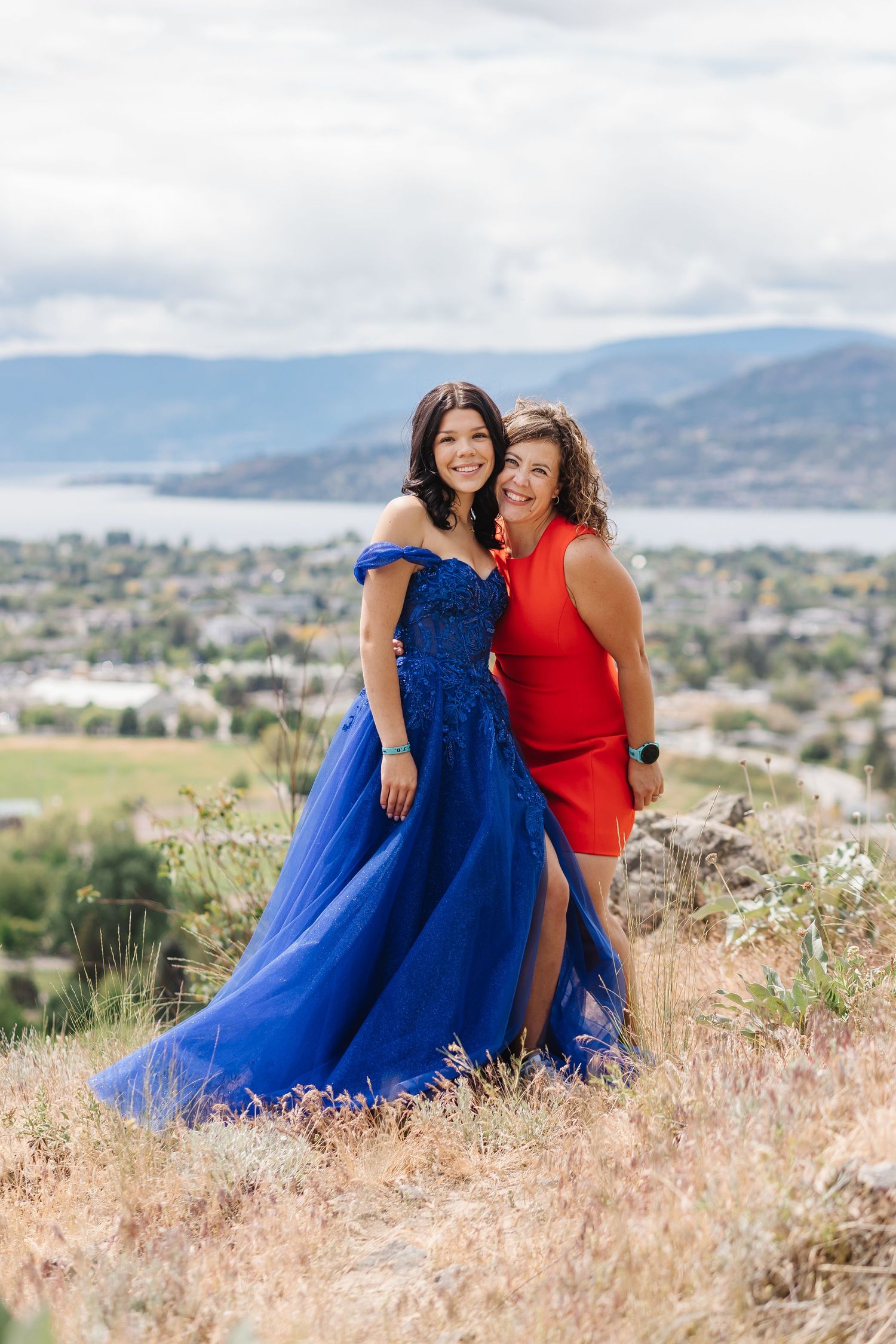 Two friends in blue and red formal dresses pose together on a hilltop overlooking a scenic mountain landscape.