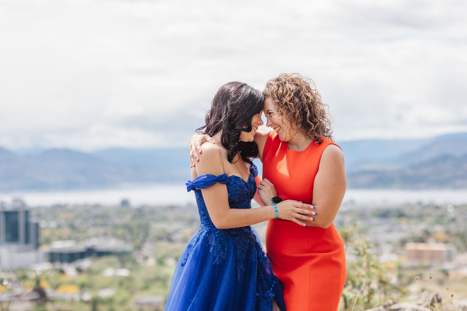 Two women in vibrant blue and red dresses share a joyful moment overlooking a scenic mountain city landscape.