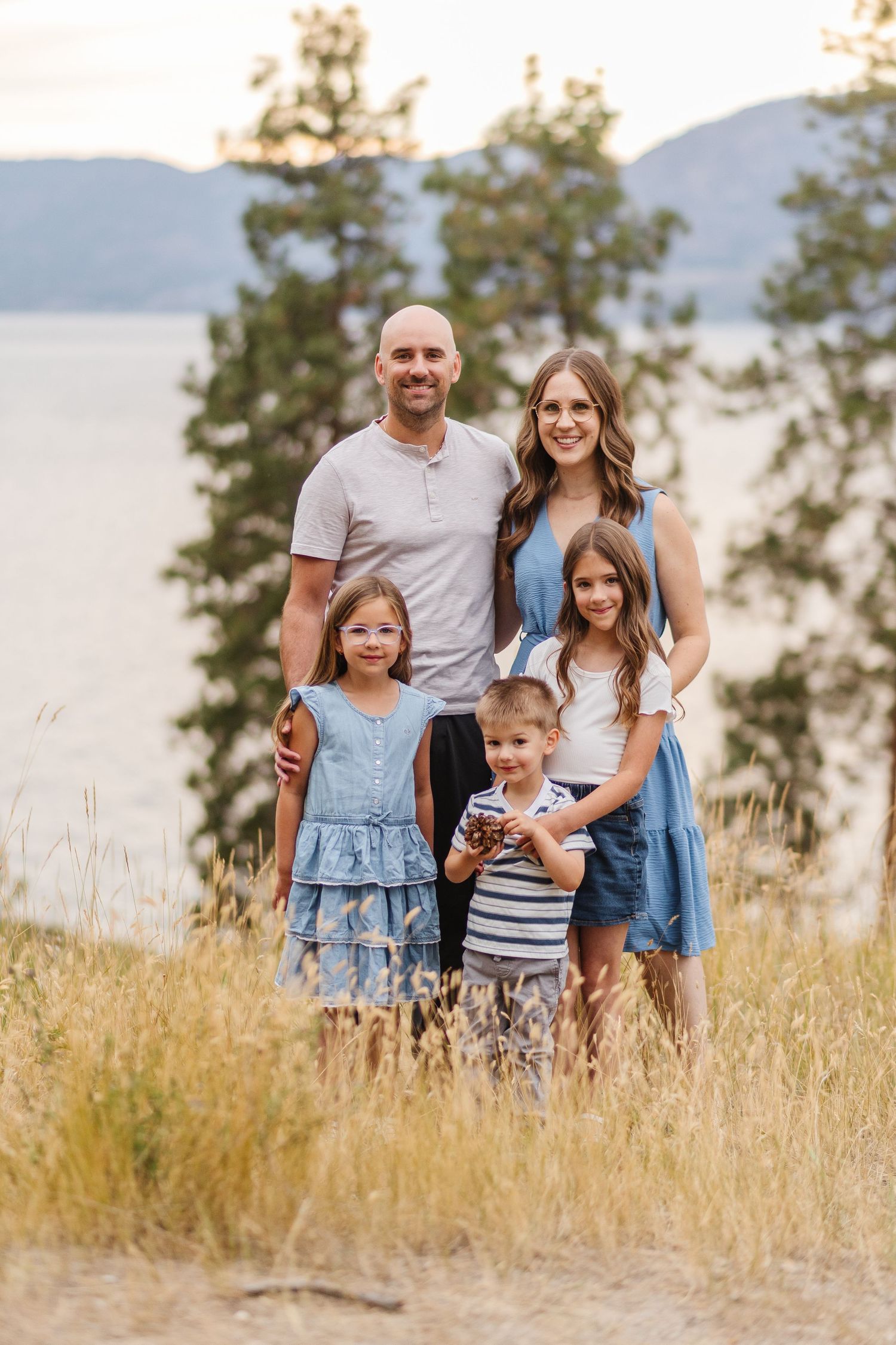 A family stands together in a golden field with mountains and trees visible in the background during sunset.