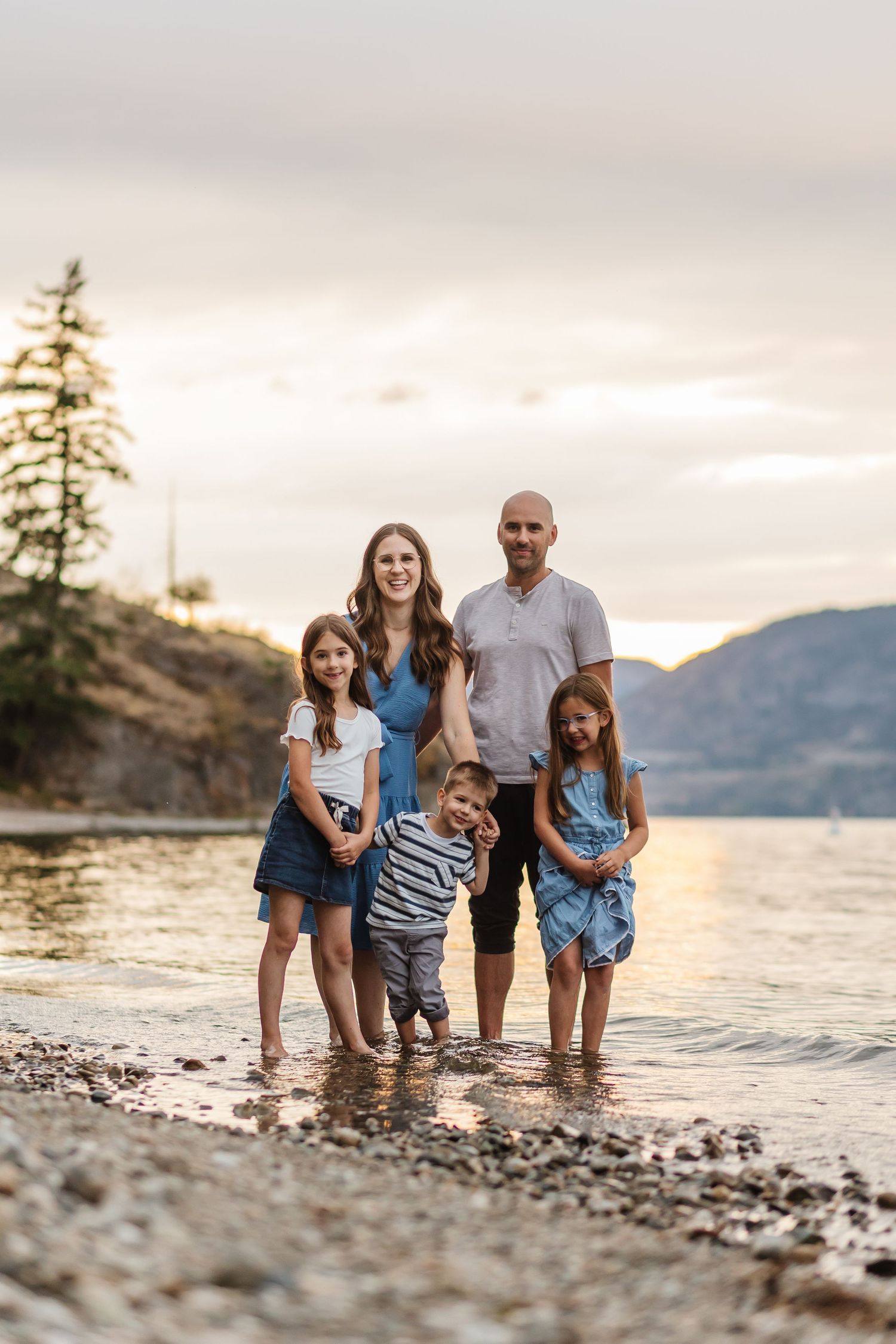A family stands together at the water's edge during sunset with mountains and trees in the background.