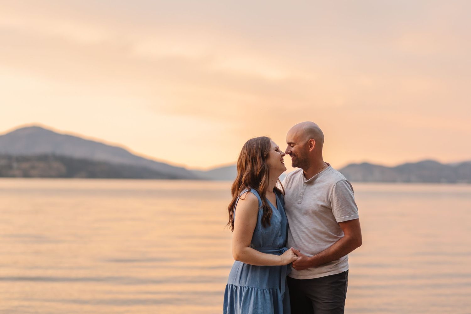 A couple shares a romantic moment at sunset by the ocean with mountains visible in the background.