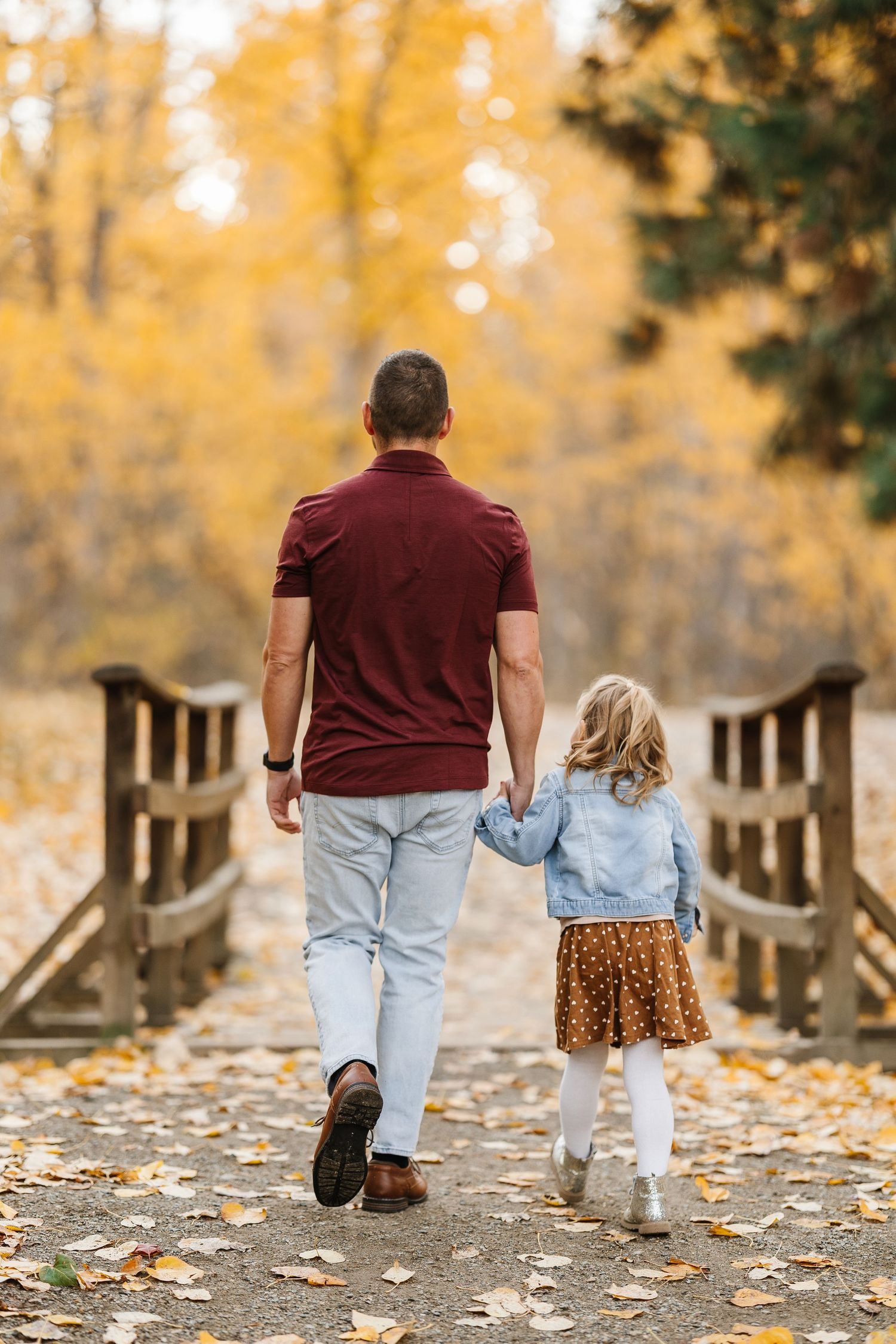 Two people walk together on a wooden bridge path covered in fallen autumn leaves with golden trees in background.