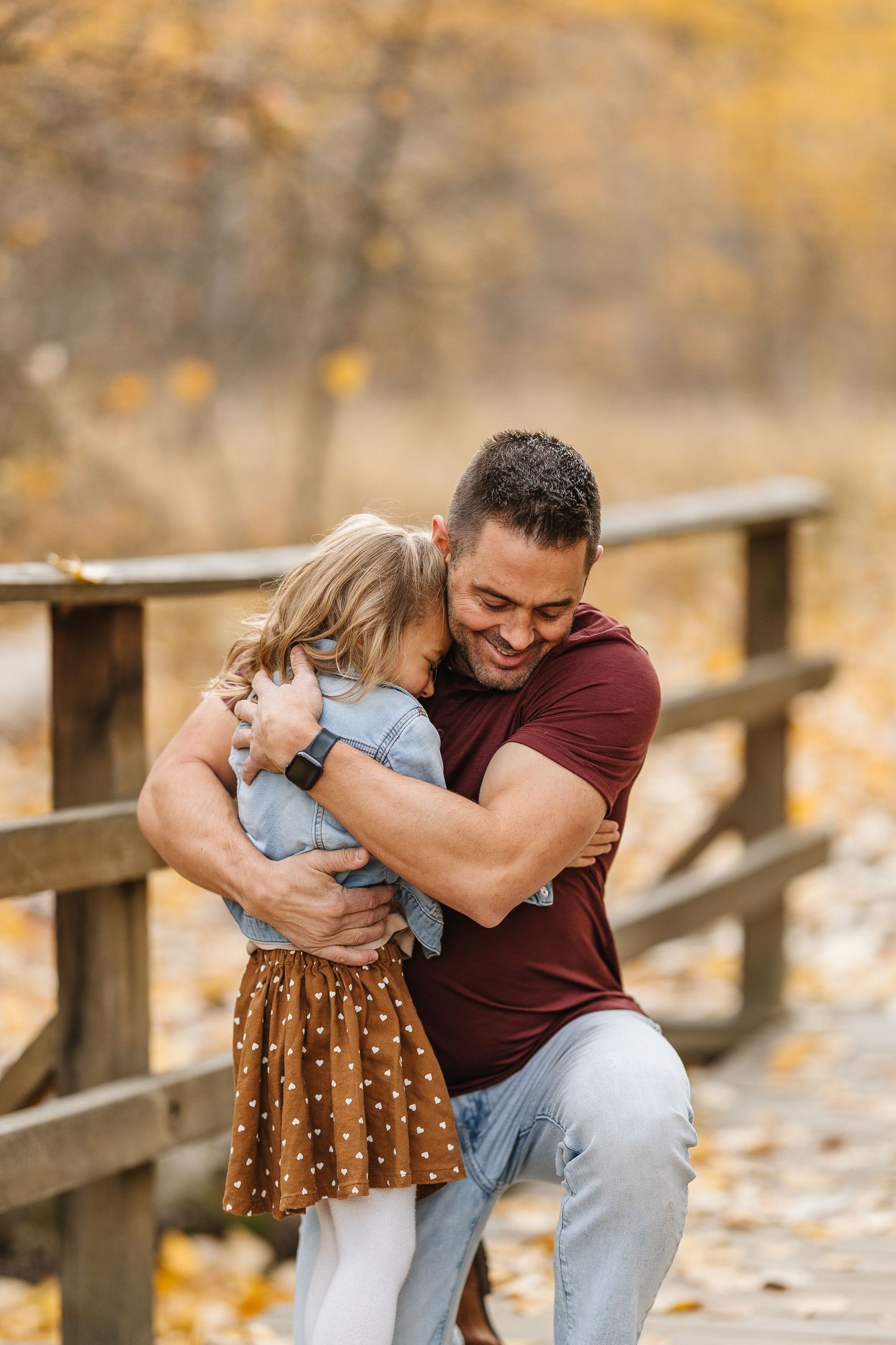 A tender embrace shared between two people on a wooden bridge during autumn with golden foliage in the background.