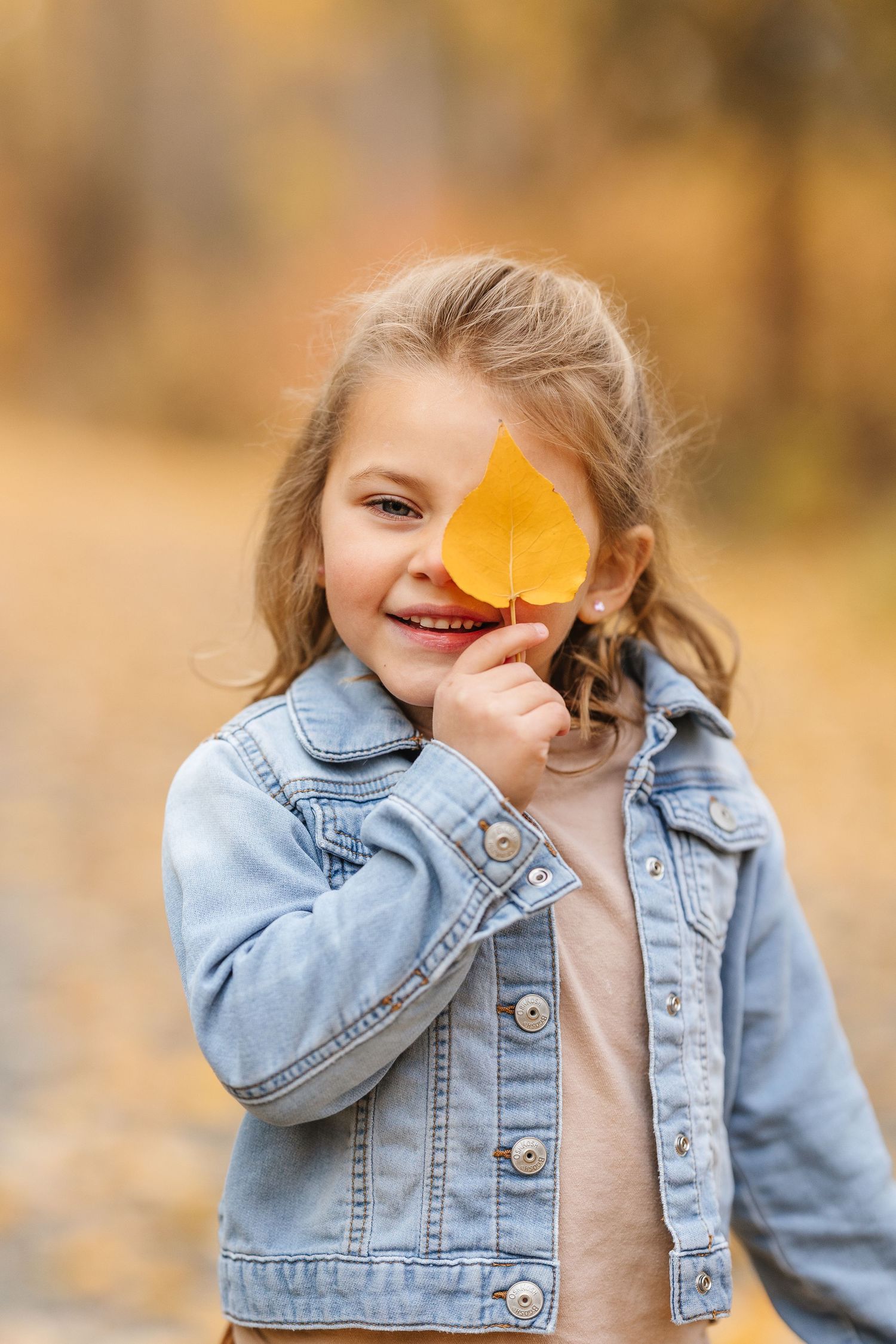 Child wearing denim jacket holds yellow autumn leaf over eye while playing outdoors in fall season.