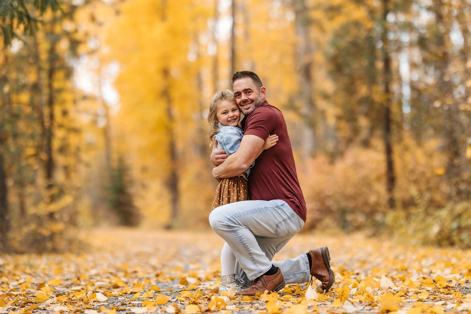 A romantic couple embraces in a golden autumn forest surrounded by fallen yellow leaves on the ground.