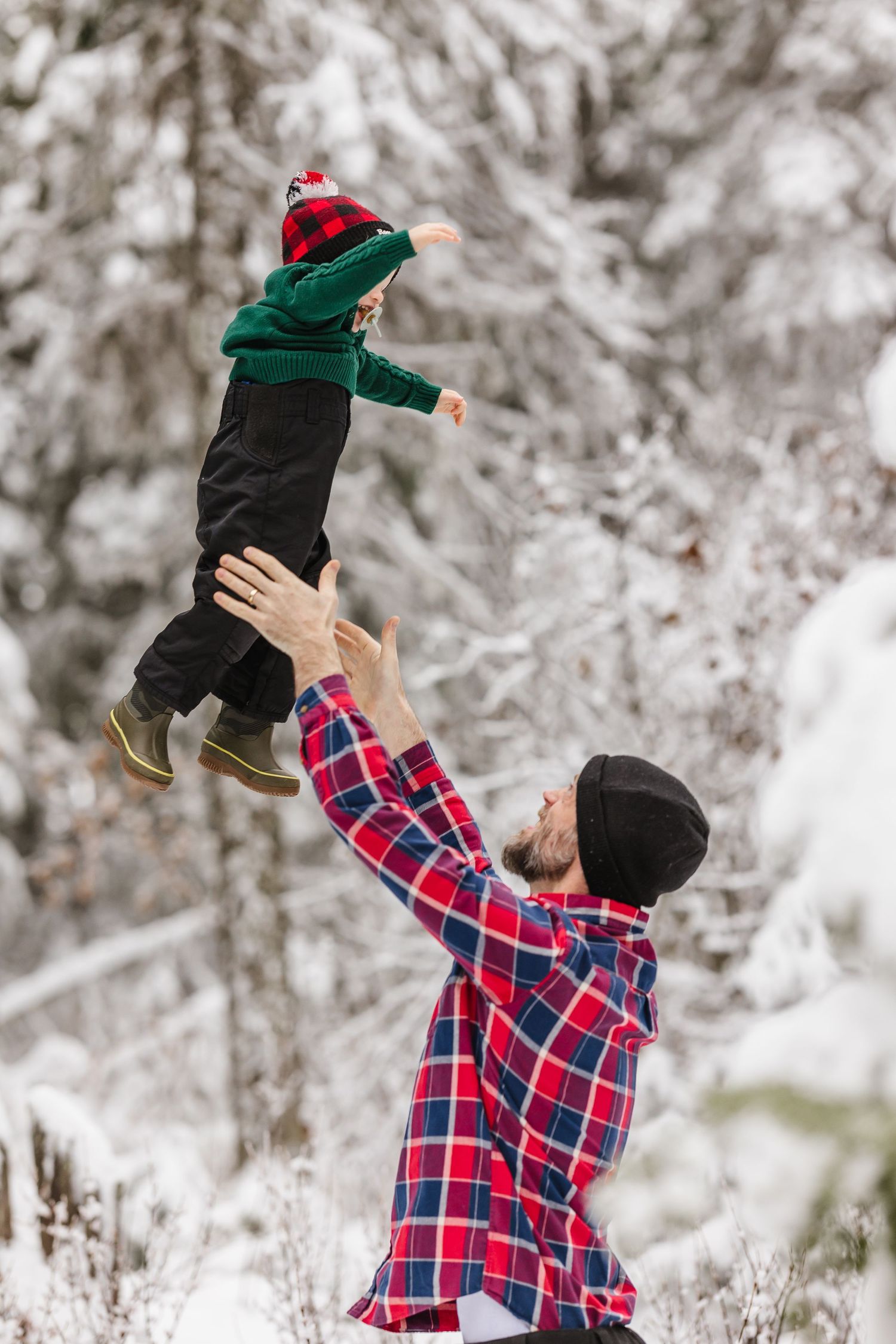 Person in plaid shirt lifts small child wearing green coat and red hat in snowy winter scene.