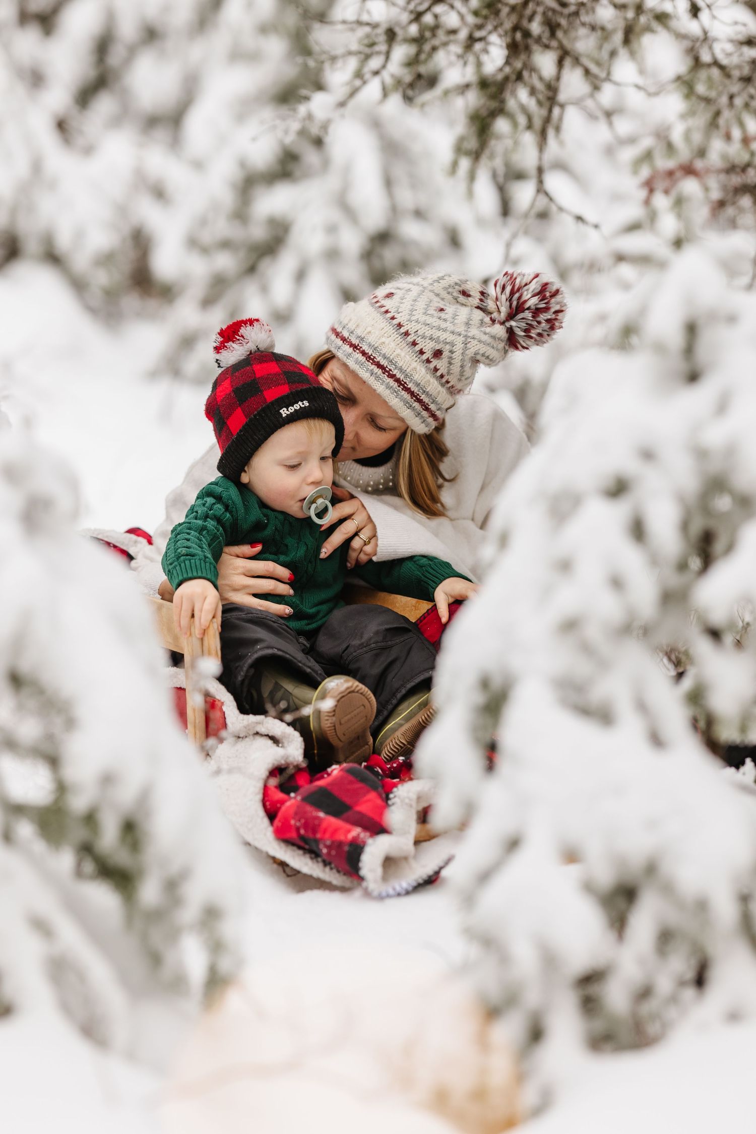 Two people wearing cozy winter knits and plaid share a tender moment in a snowy forest setting.