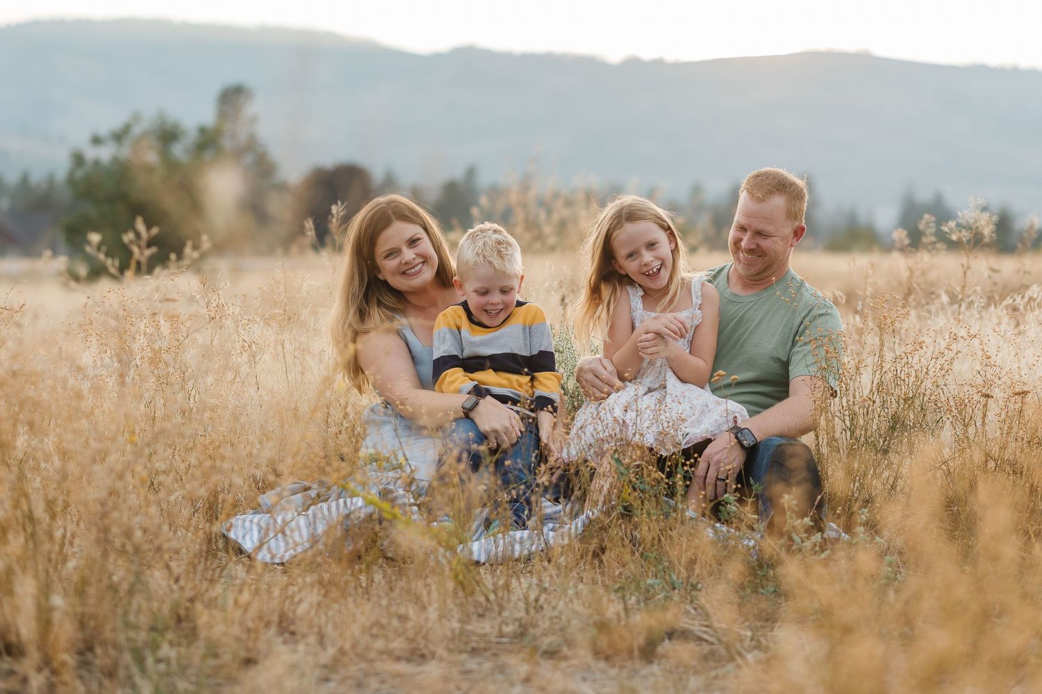 A happy family sits together in a golden wheat field at sunset with mountains in the background.