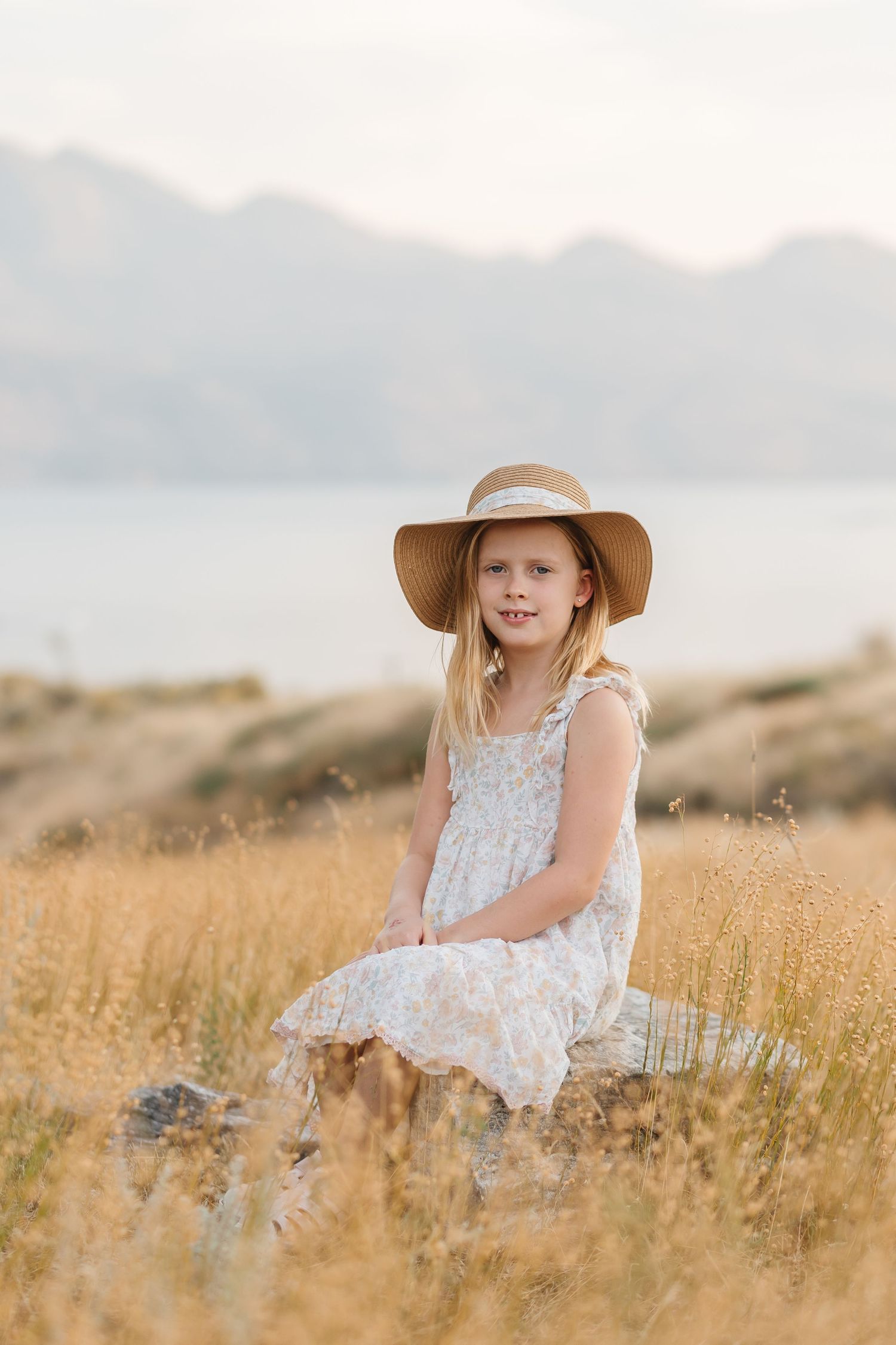 A person in a white dress and floppy sun hat sits among tall golden grass at sunset in a dreamy outdoor setting.